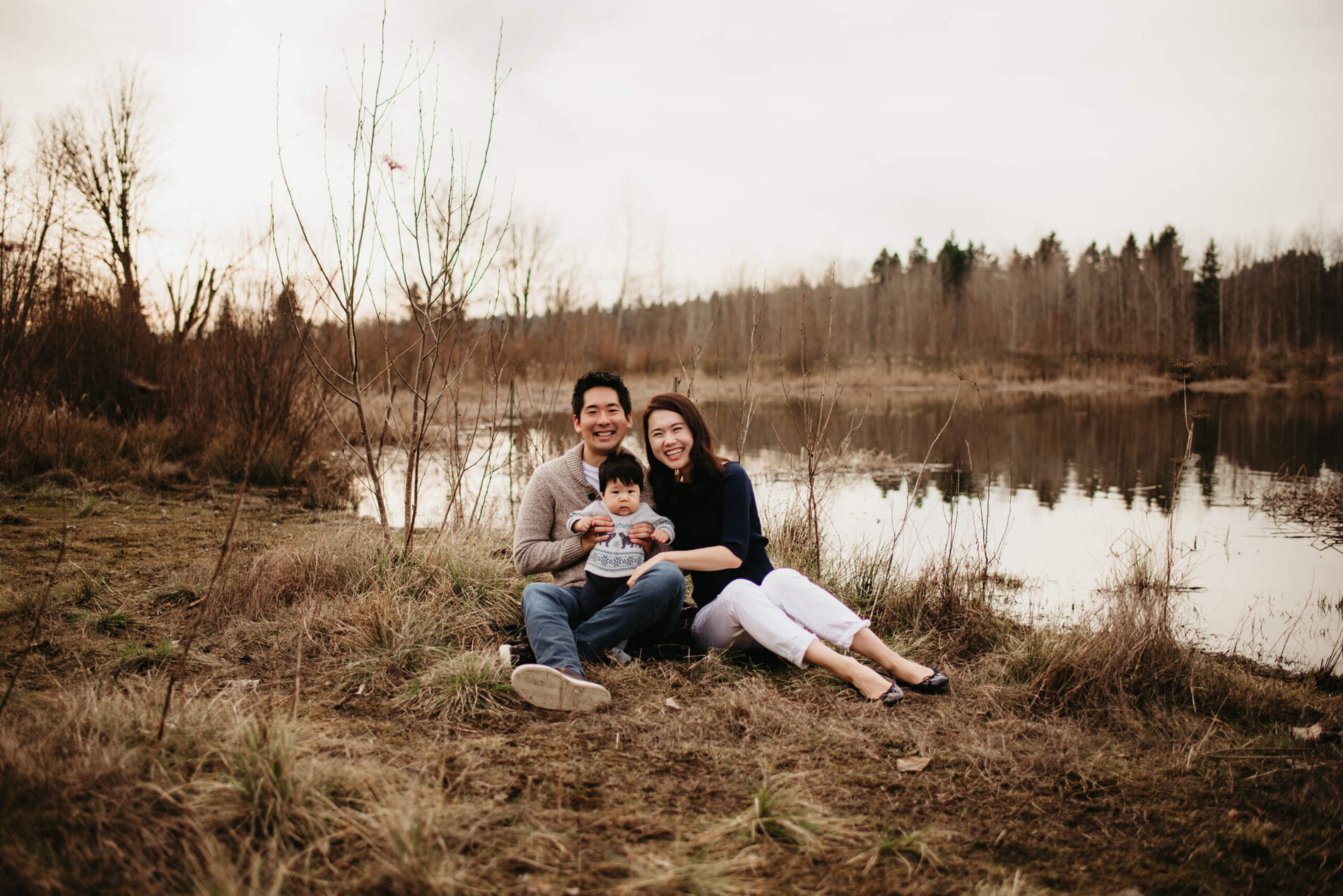 Family portrait of young parents sitting with their toddler son in a field and pond in the background