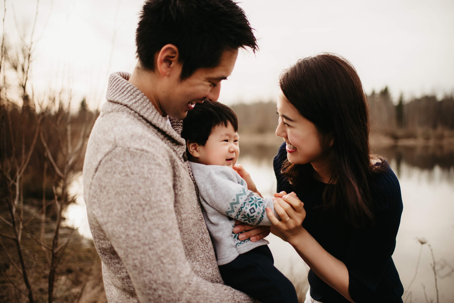 Family lifestyle photo session of two happy parents playing with their toddler son in a natural setting with a pond in the background