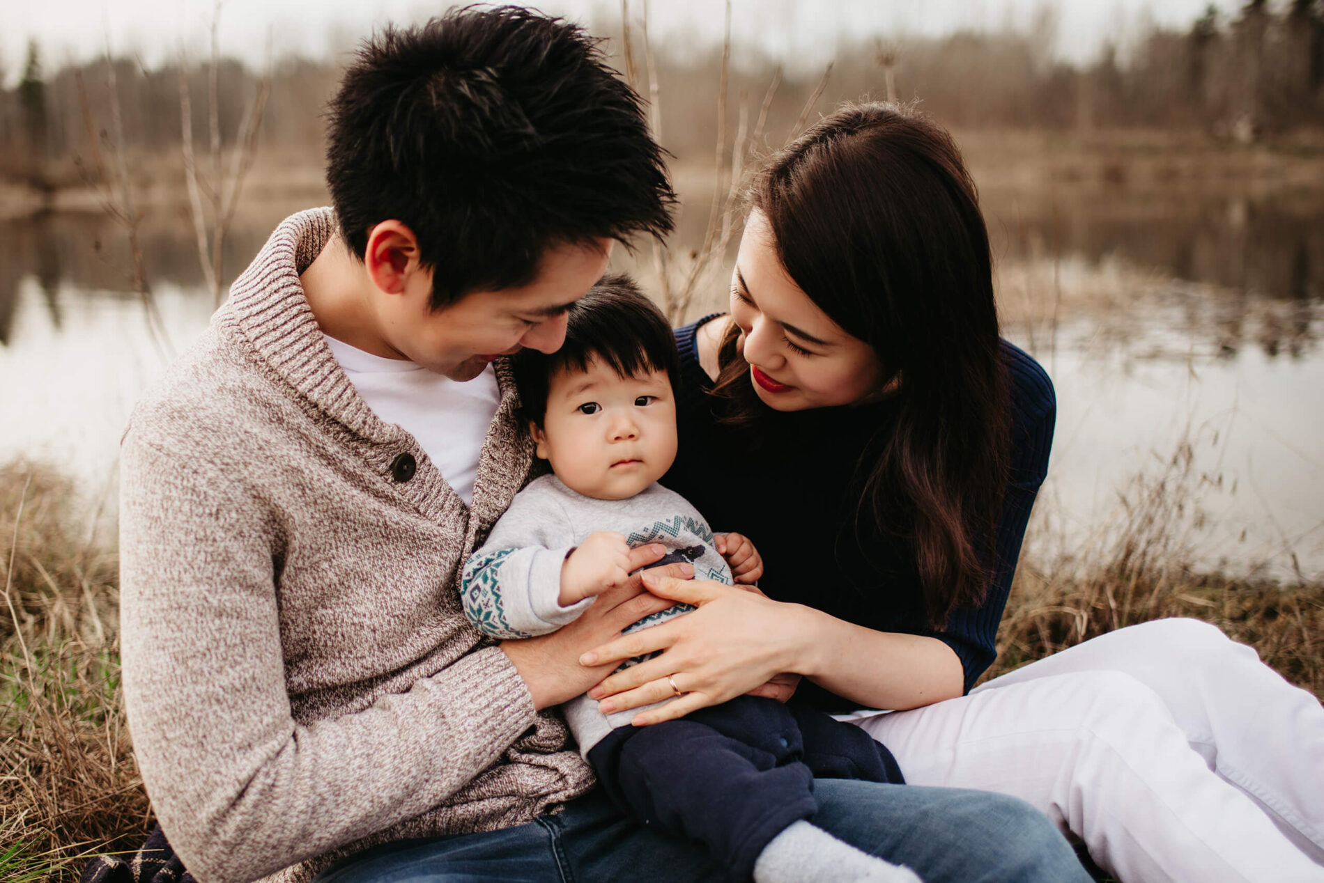 Family lifestyle photo session of two happy parents playing with their toddler son in a natural setting with a pond in the background
