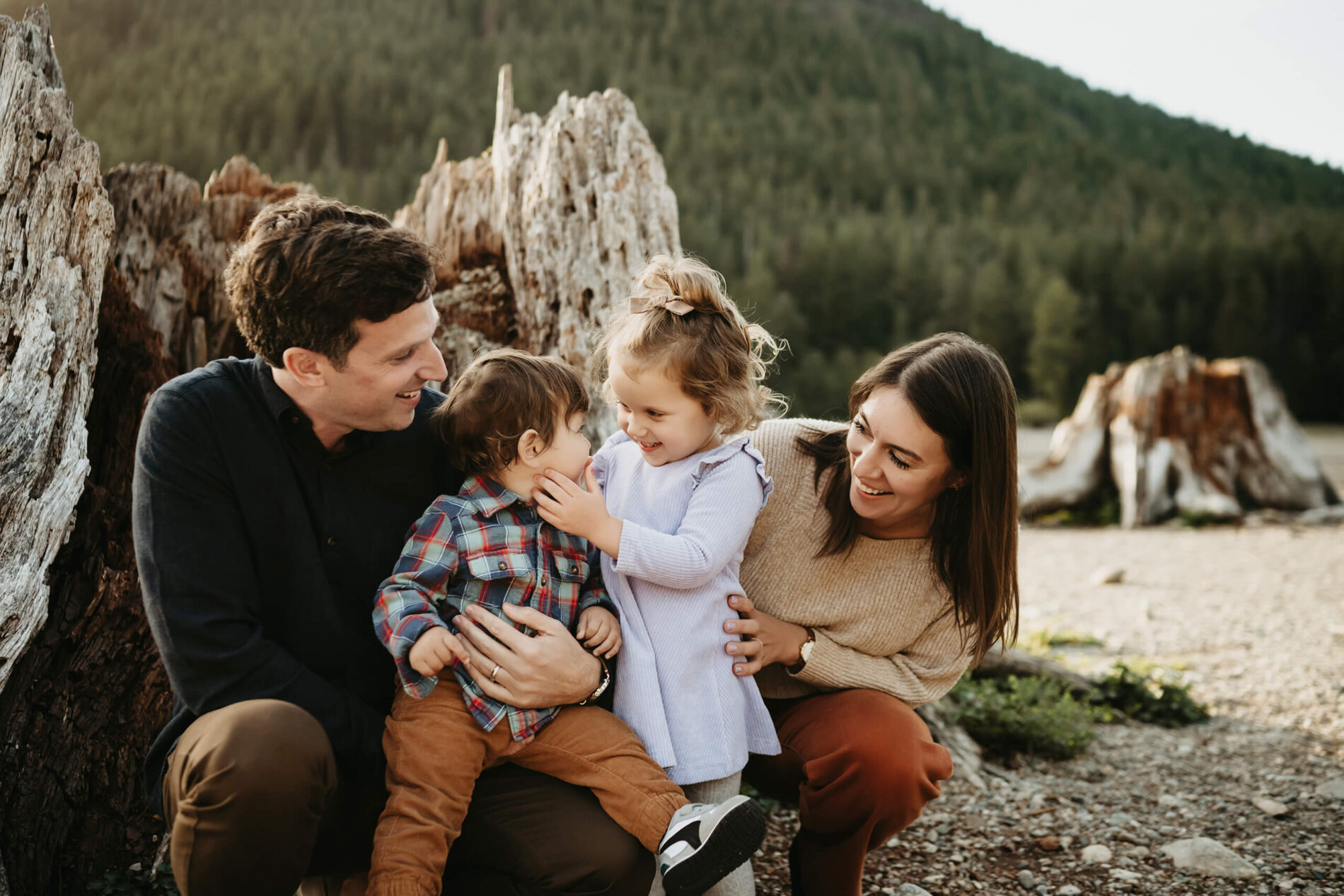 Playful Seattle family photo by Lana Sky Photography: mom, dad, toddler daughter and baby son laugh next to a weathered stump at Rattlesnake Lake with evergreen mountain backdrop.