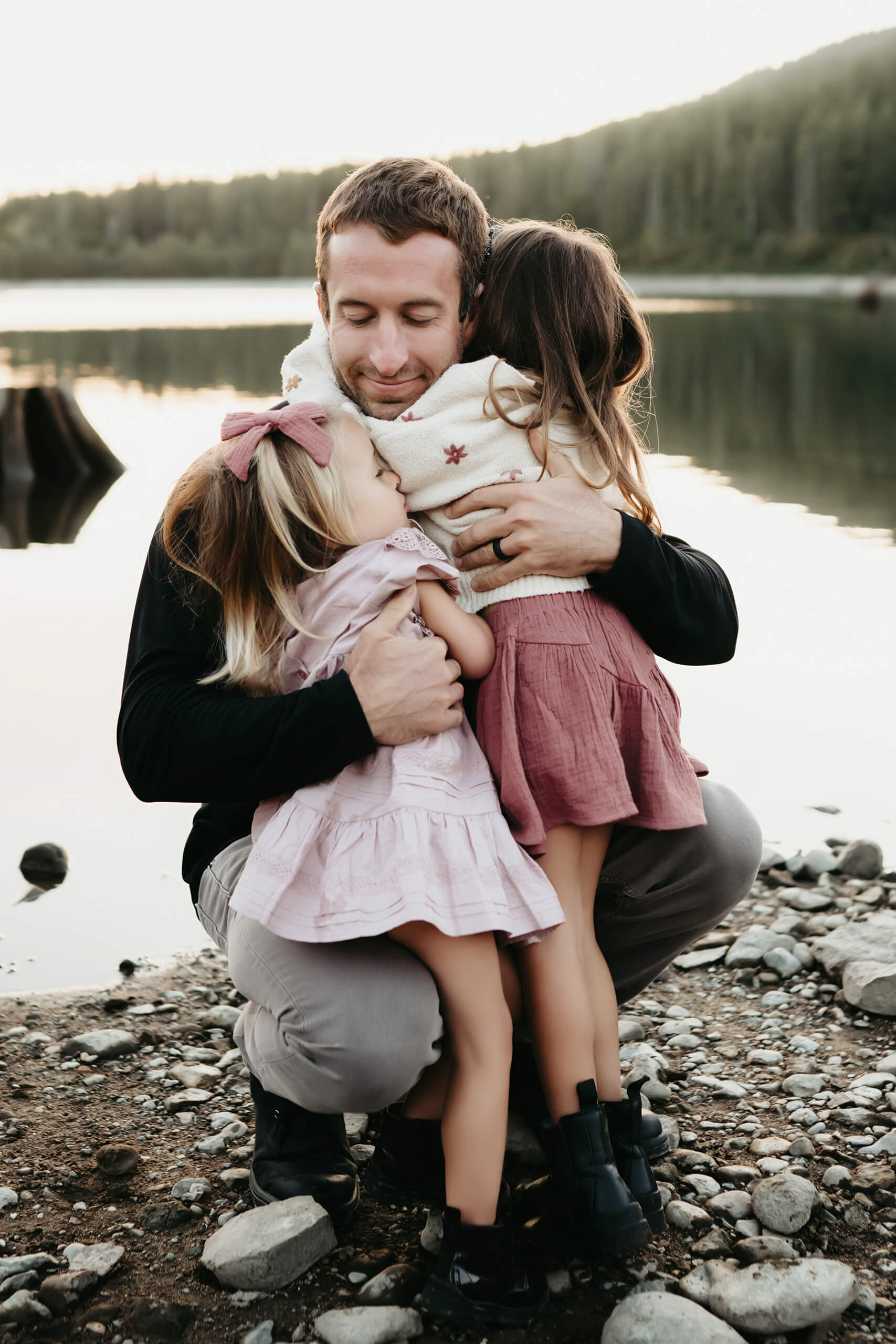 A family portrait of dad hugging his two daughters at a beach with lake and mountains in the background