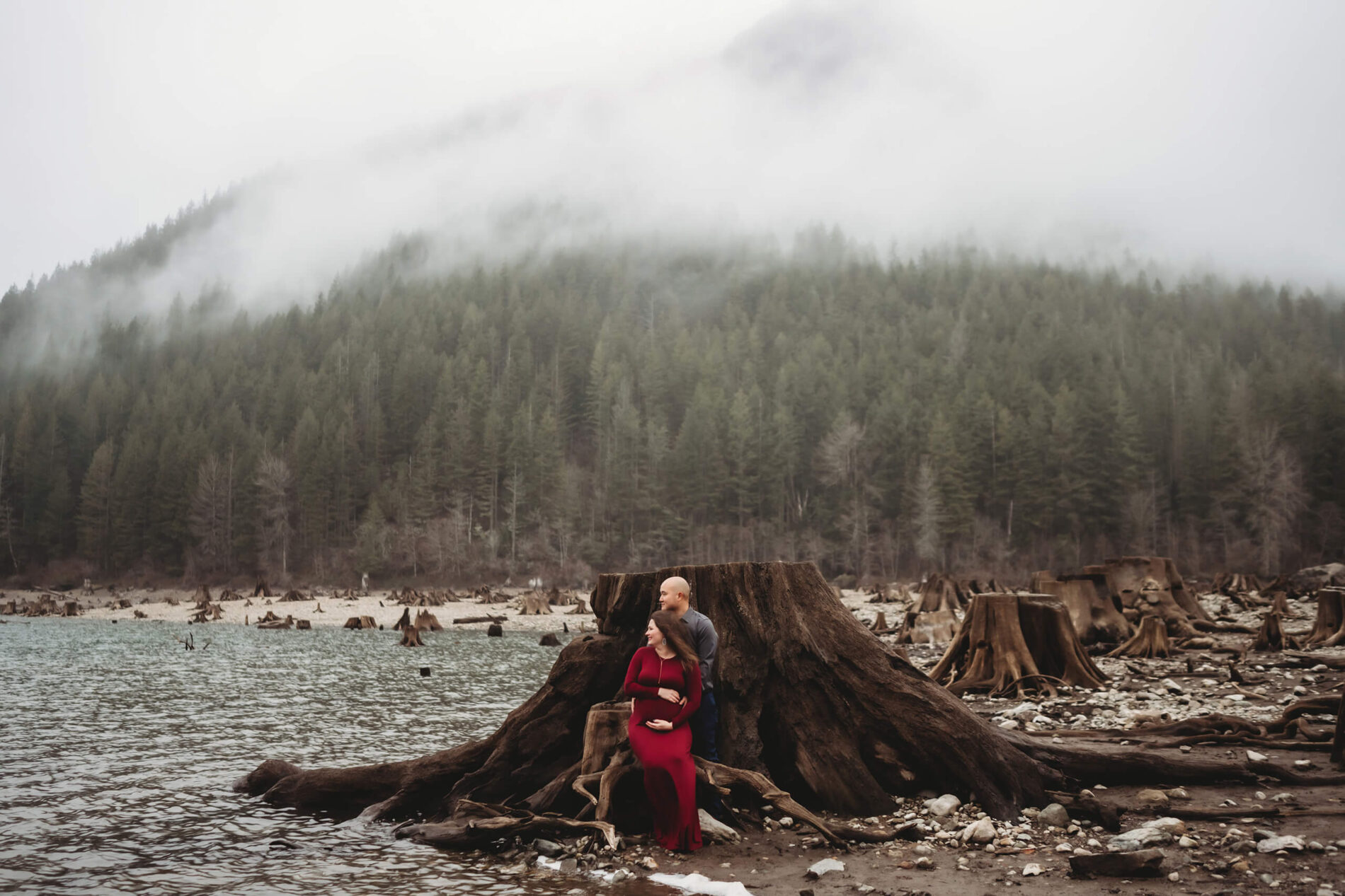A couple posing during an outdoor winter maternity shoot with lake, trees and mountains in the background