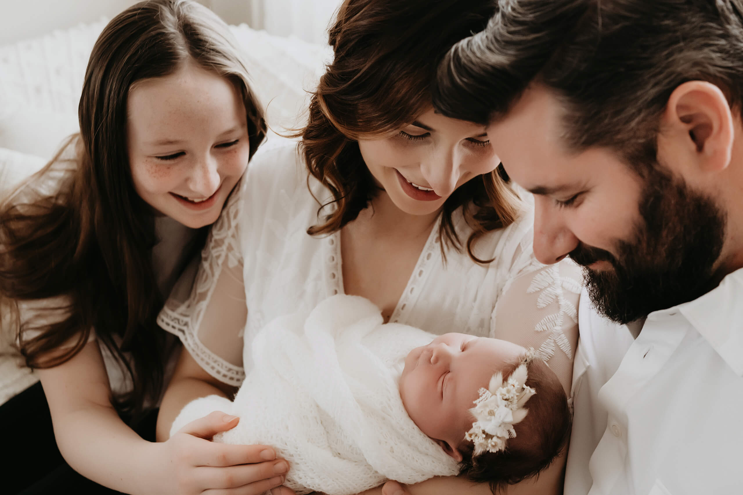 Seattle newborn photographer captures tender family moment in Kirkland studio: mom cradles swaddled baby girl with floral headband as dad and big sister smile in soft window light.