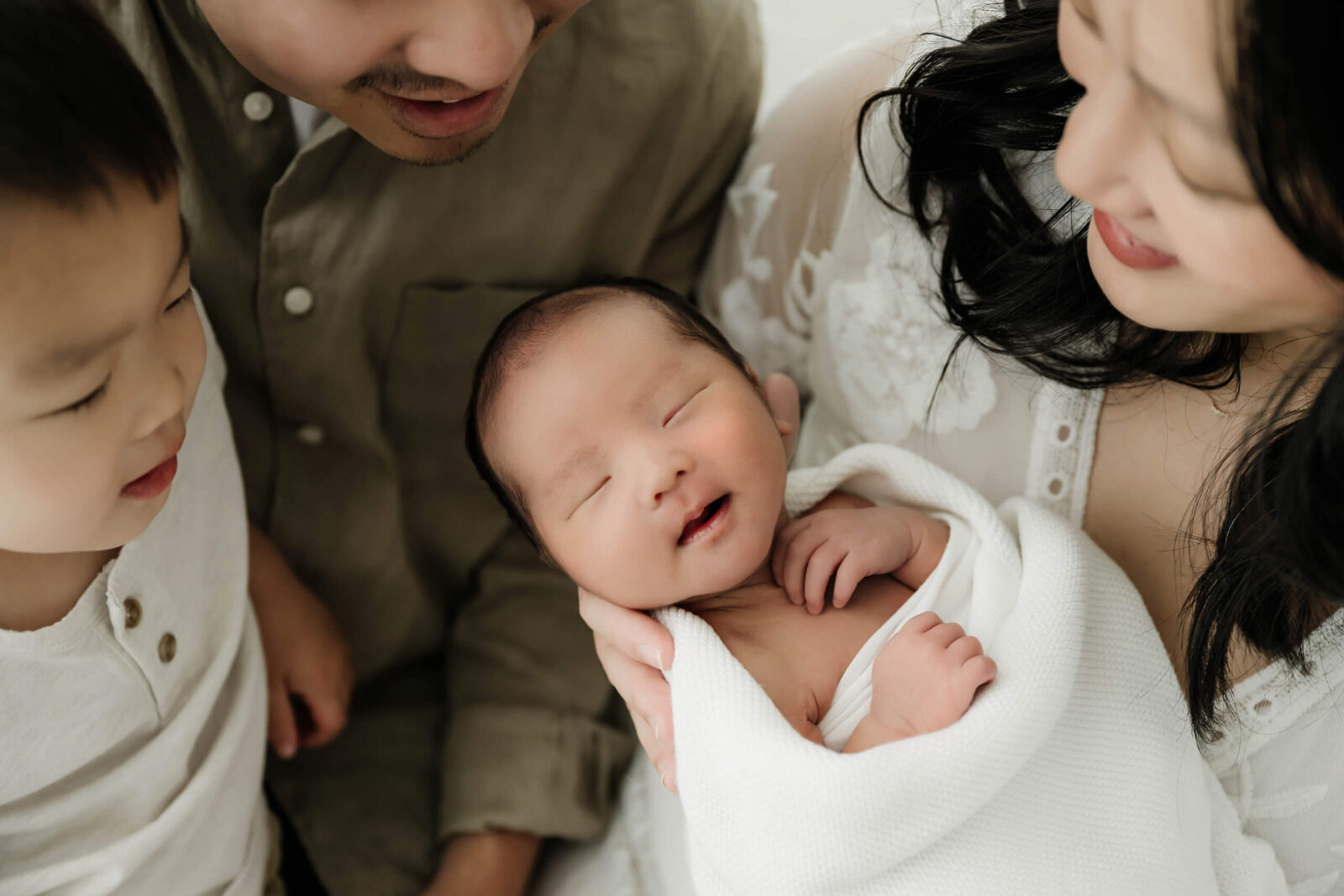 Parents and toddler sibling admire their newborn during a Seattle newborn family photoshoot, baby swaddled in white with a peaceful smile