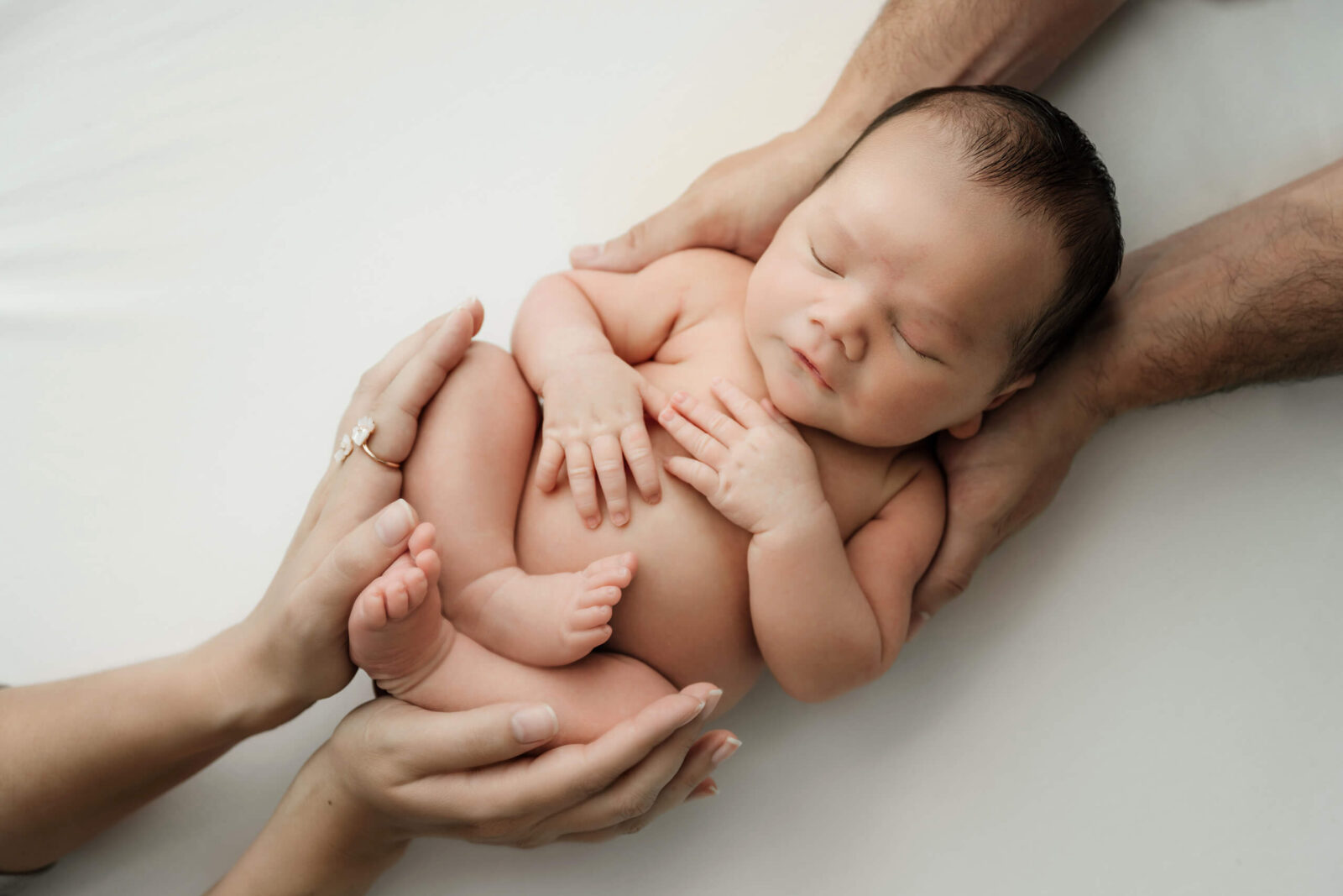 Tiny newborn rests curled on a white backdrop, gently supported by parents’ hands during a Seattle newborn family photoshoot in studio