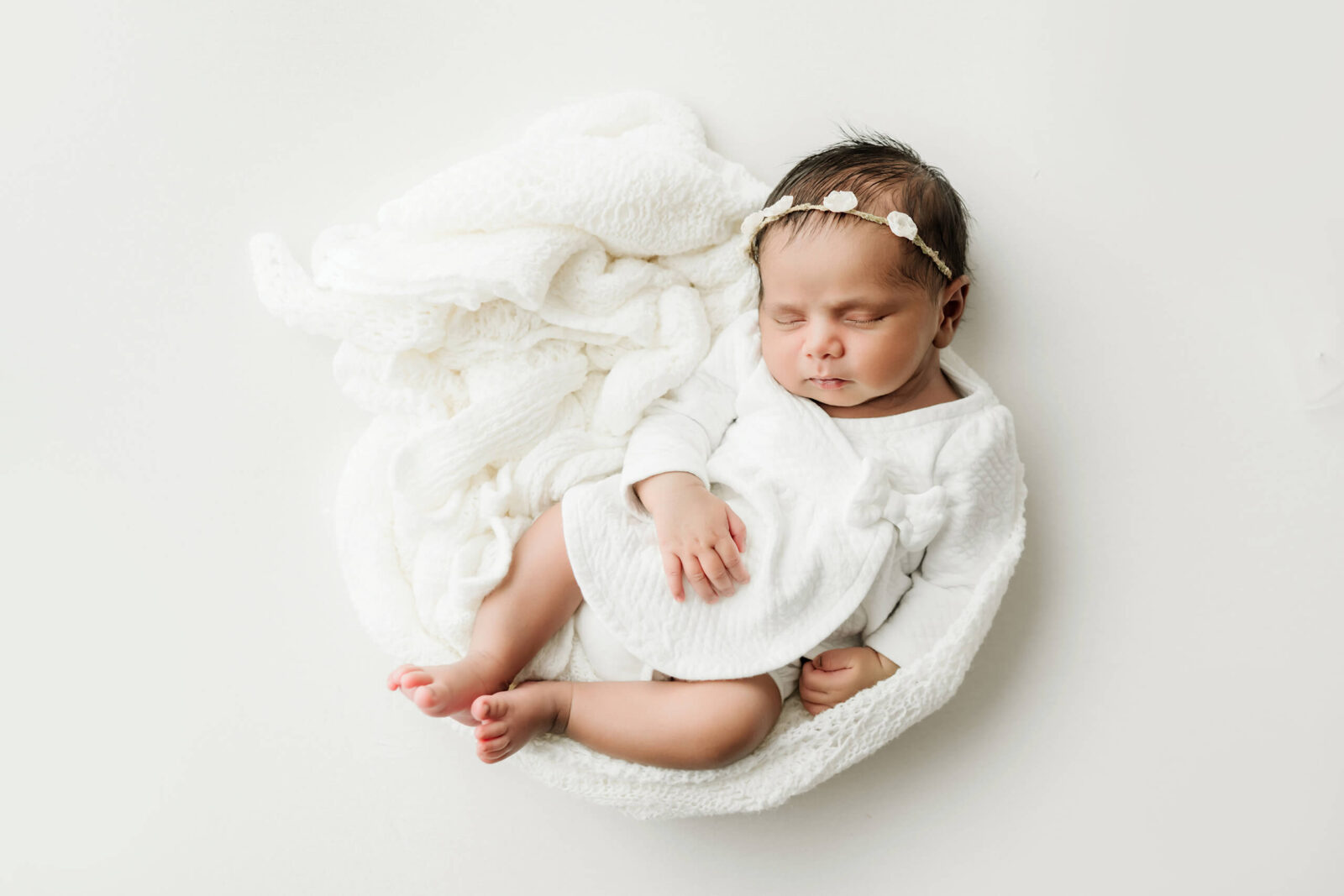 Sleeping newborn in an all-white outfit with a floral headband, posed on a soft knit layer for a Seattle newborn family photography studio session