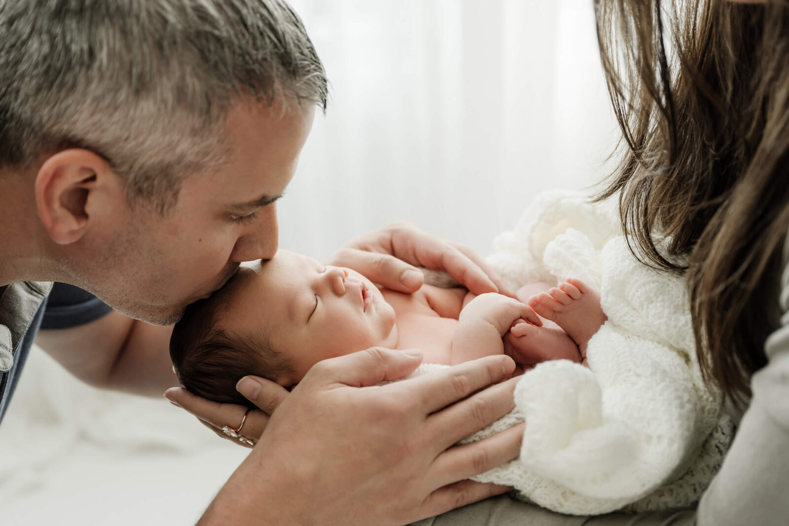 Dad kisses newborn’s forehead as parents hold baby close during a Seattle newborn family photoshoot, soft knit wrap and gentle connection