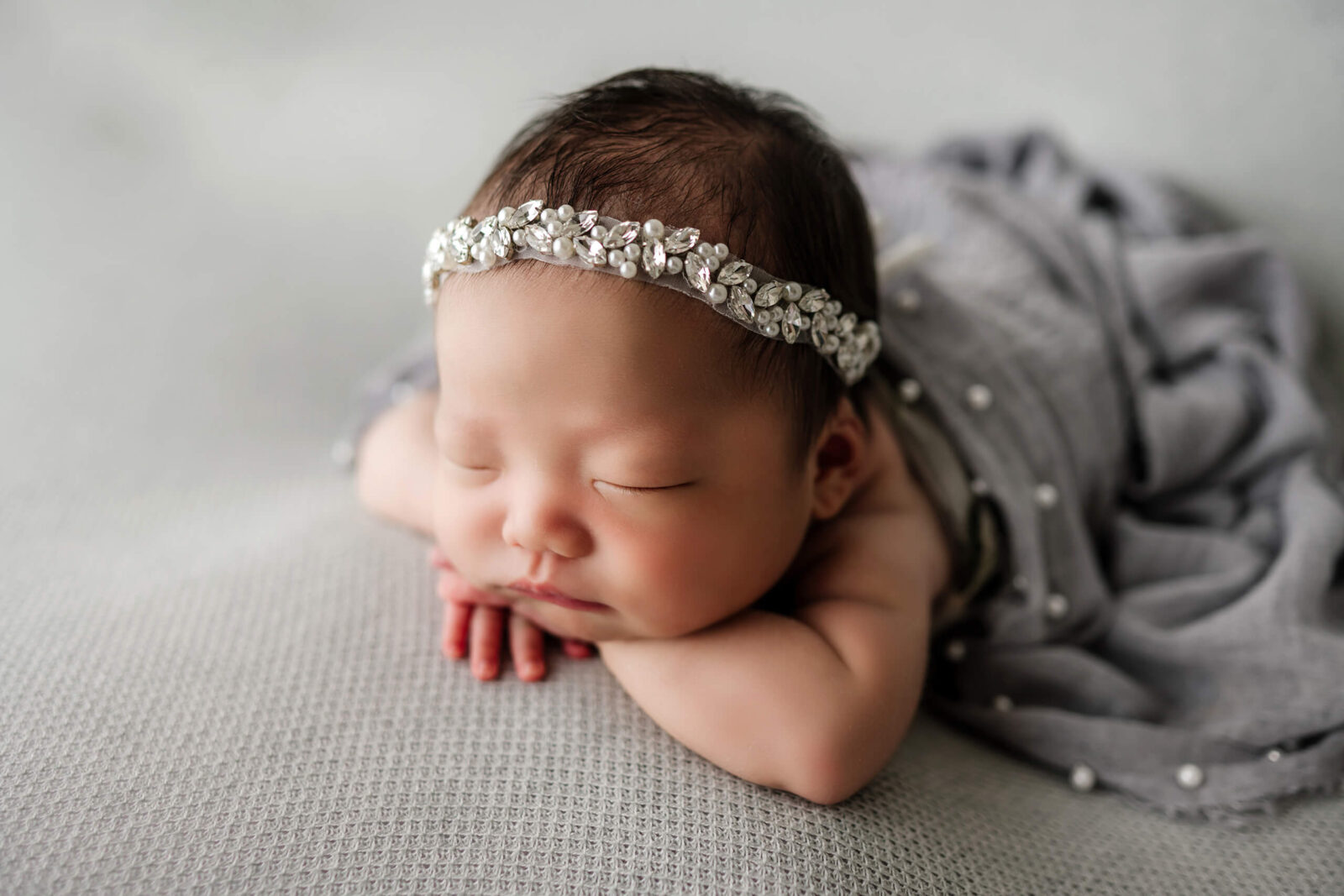 Seattle newborn studio portrait: sleeping baby girl in jeweled pearl headband rests chin on hands, tummy-down on soft gray textured blanket with matching beaded wrap—Lana Sky Photography.