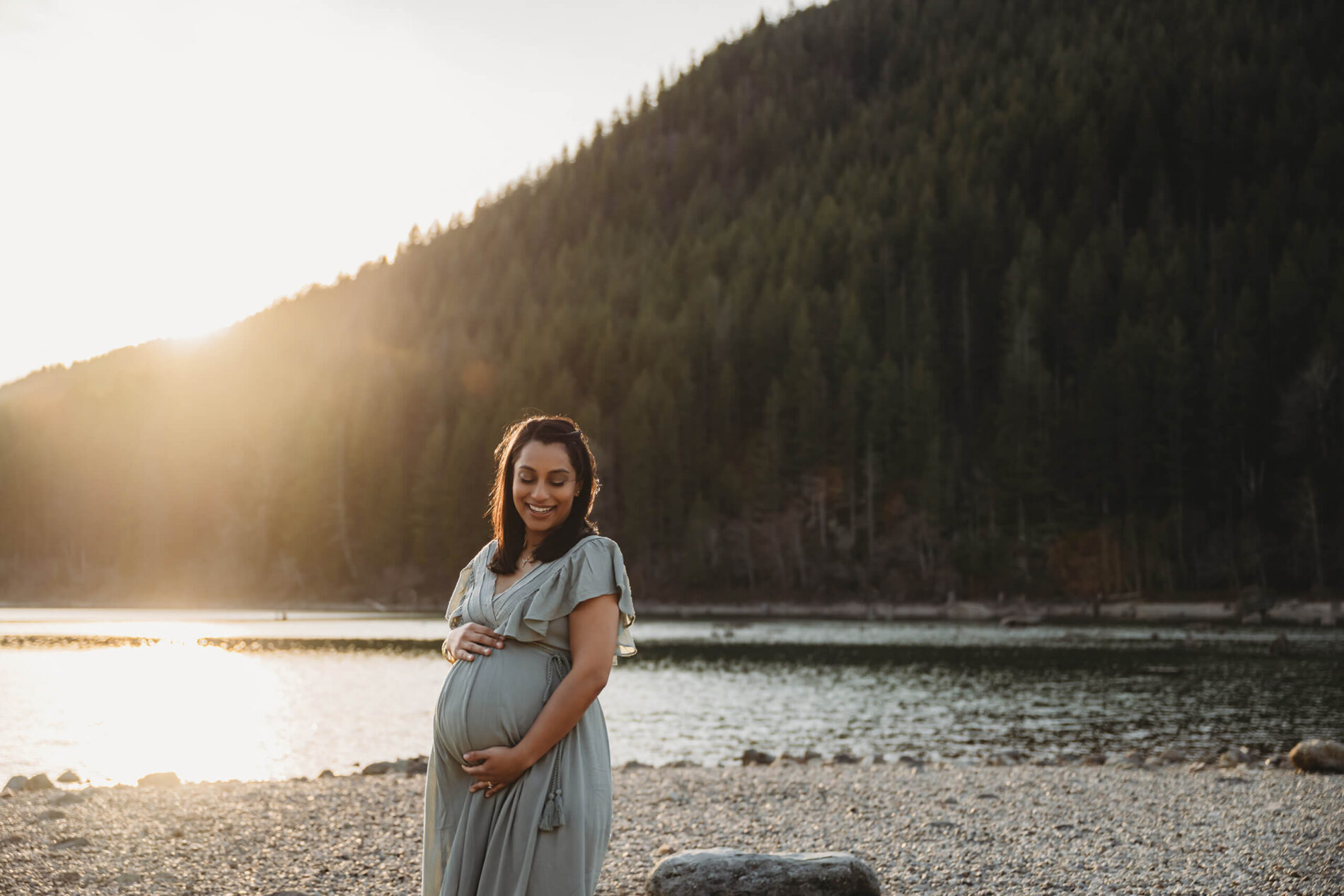 Young smiling woman during a winter maternity photo shoot with lake and mountain in the background during sunset