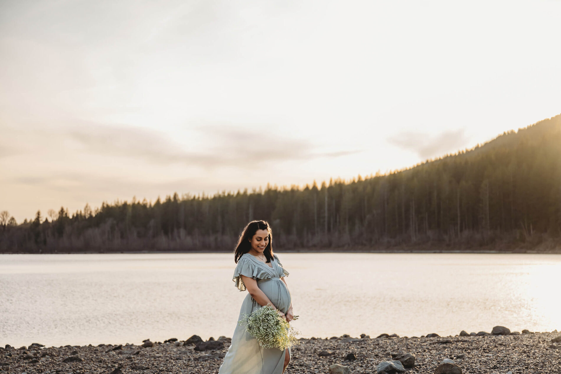 A young woman posing during an outdoor winter maternity photo shoot with beautiful lake and mountain in the background during sunset