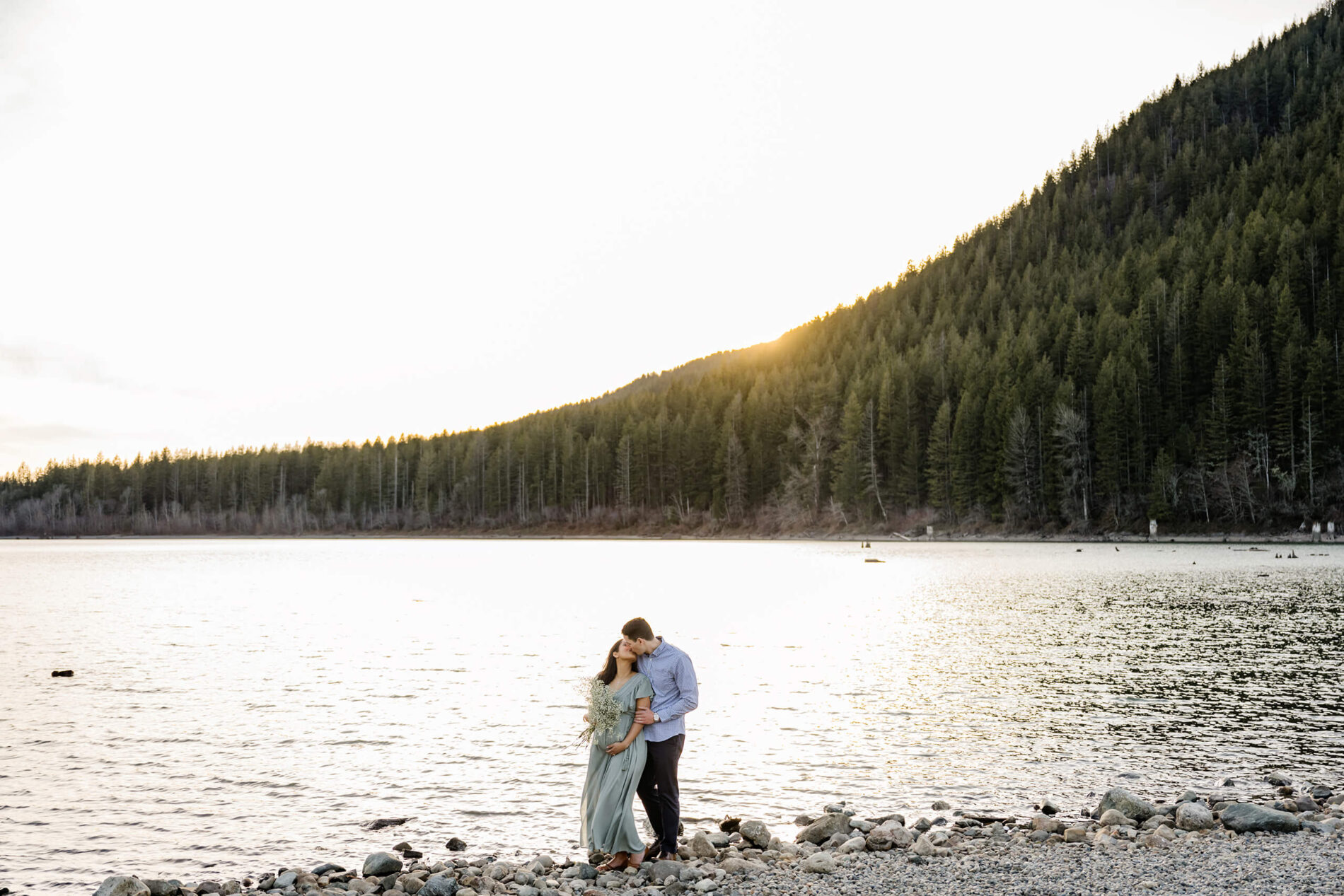 A young couple kissing during an outdoor winter maternity photo shoot with beautiful lake and mountain in the background
