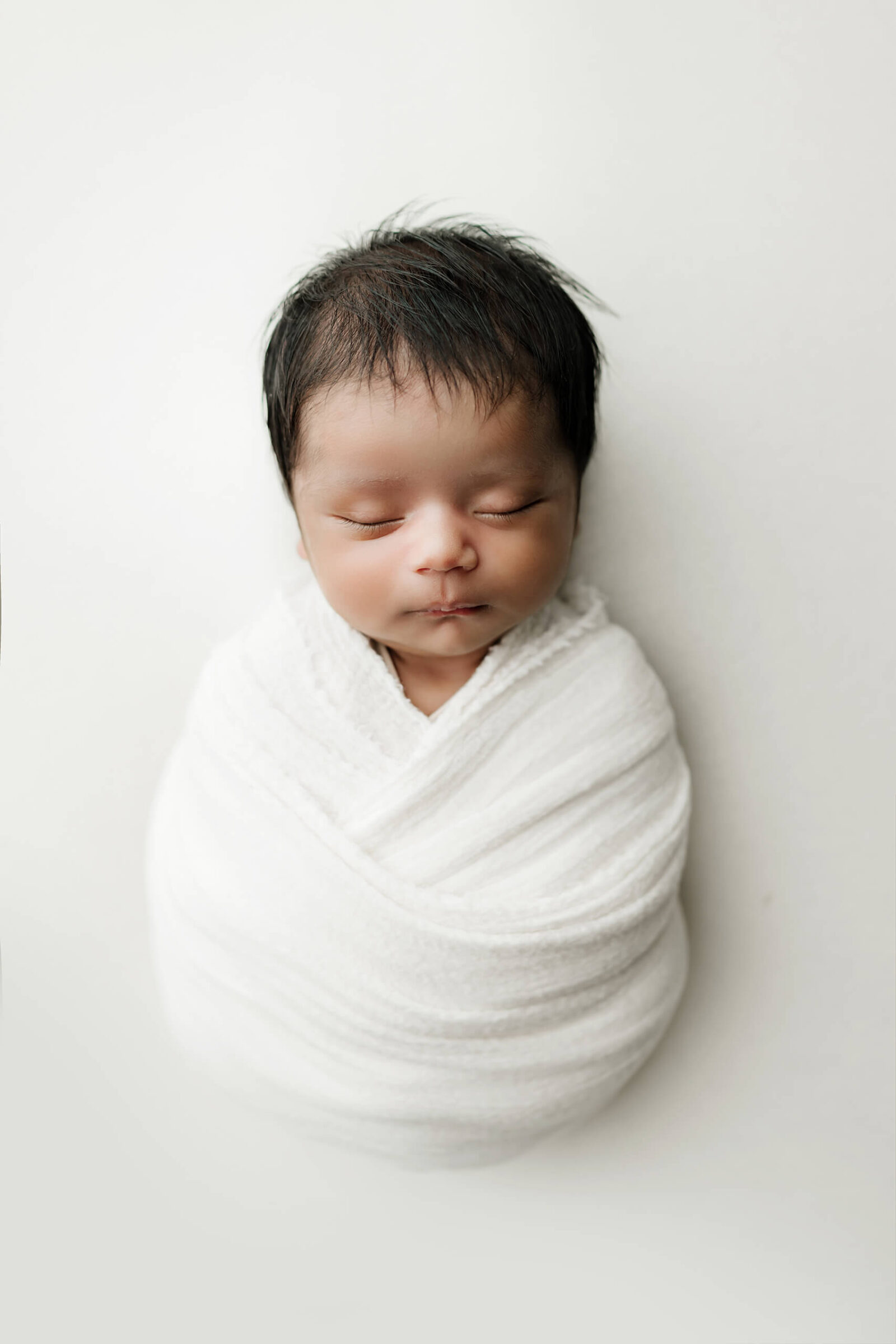 Baby boy swaddled in a white blanket against a white studio background.