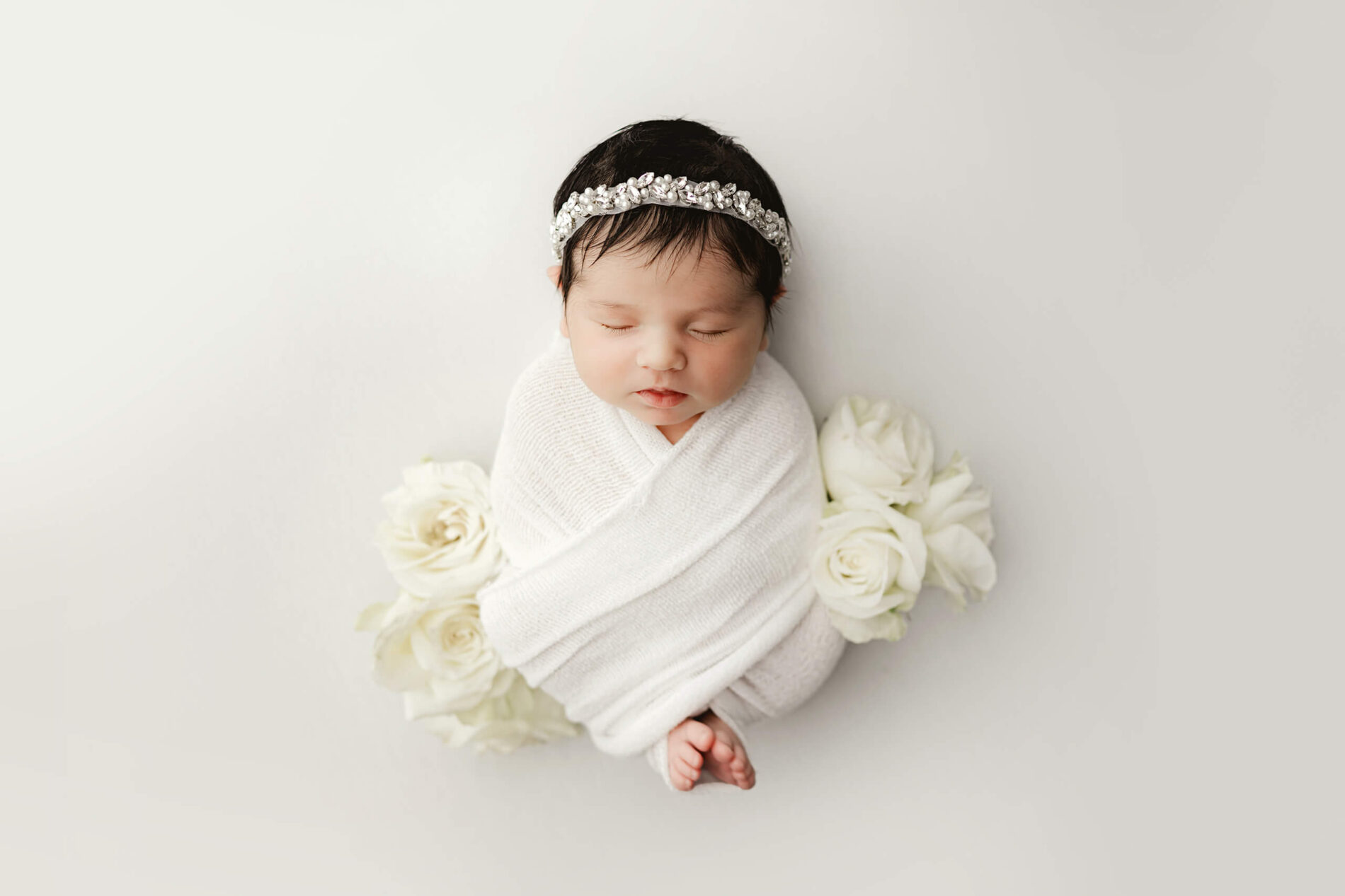 Sleeping baby girl with a beautiful headband and wrapped in blankets posed on white background with white roses during newborn photo shoot