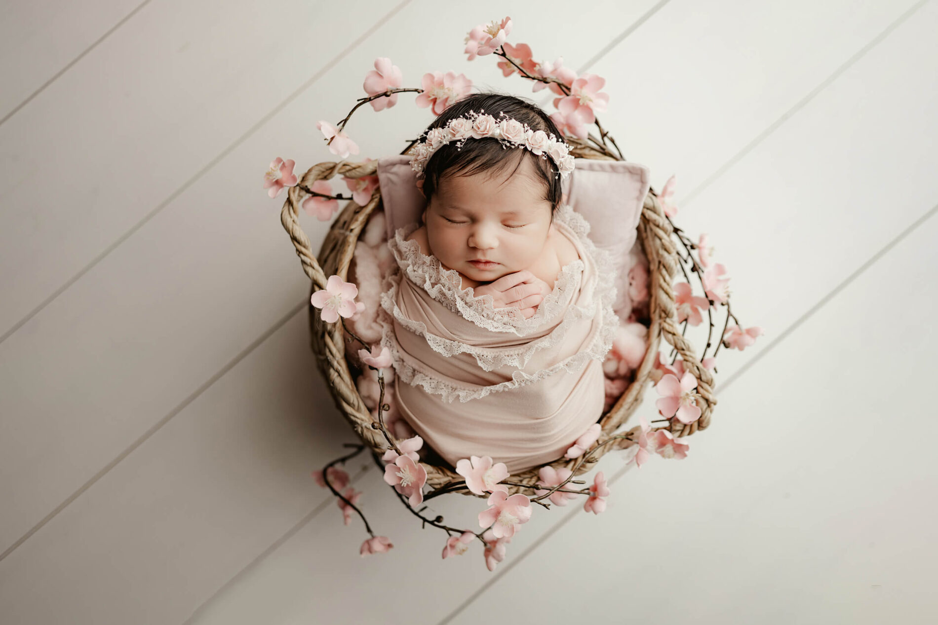 Newborn photo shoot of sleeping baby girl swaddled in pink wraps with pink floral headband posed in baskets with pink flowers
