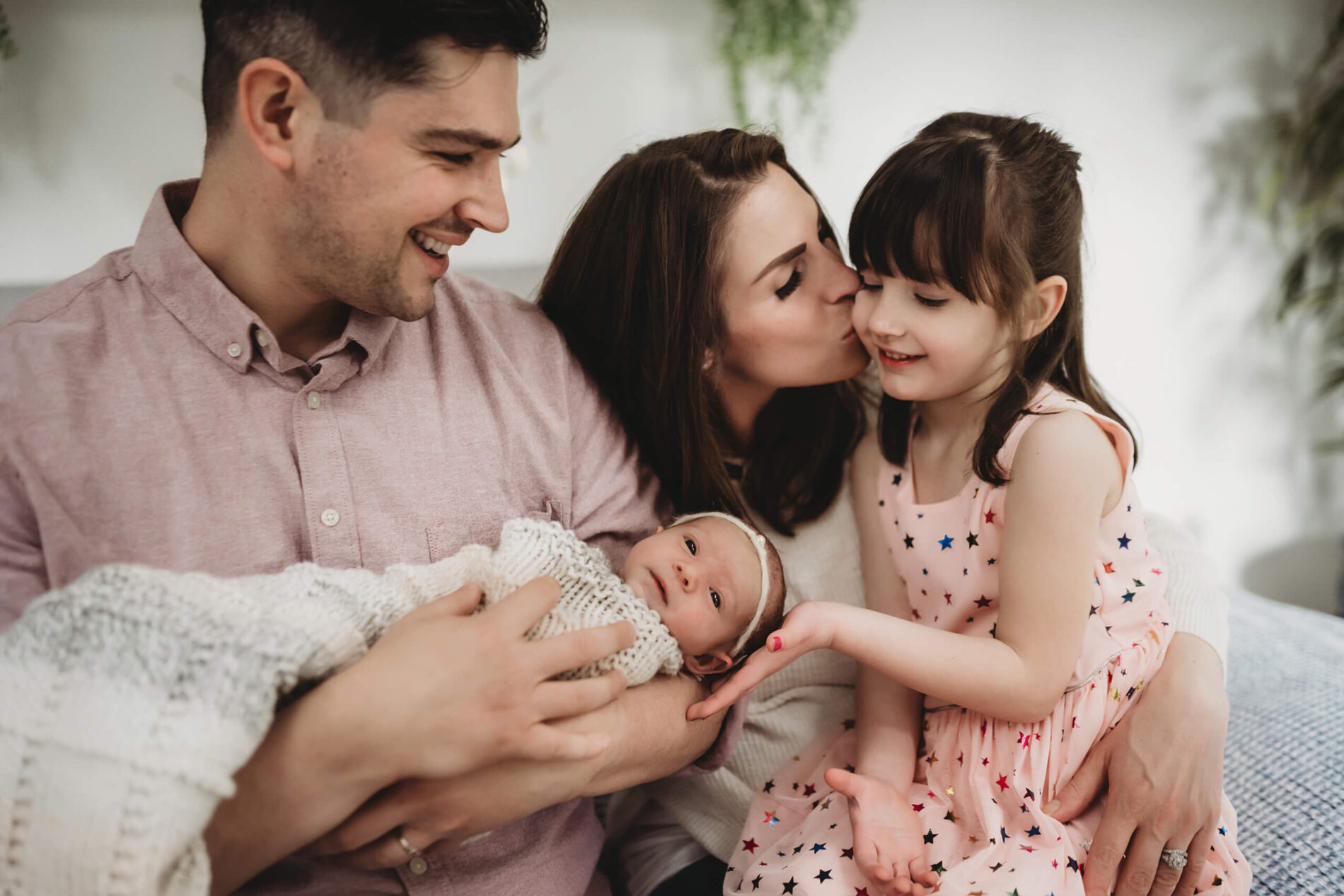 Family sitting on the bed during at home newborn photo shoot, kissing, hugging, and interacting with one another