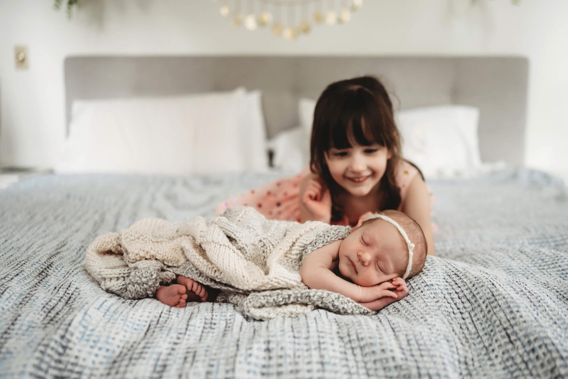 Siblings sitting on the bed during at home newborn photo shoot, older sister admiring baby sister