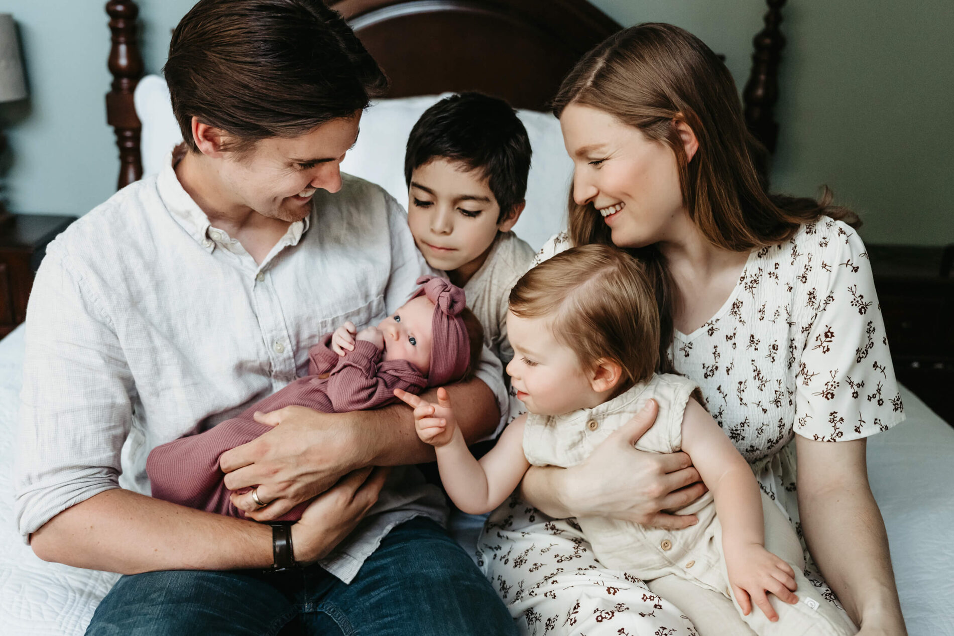 Parents and older siblings posing on a bed with baby girl during a newborn photo shoot at home, with everyone looking at the baby