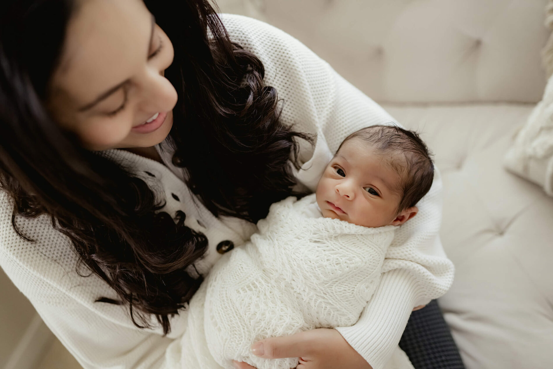Moment during newborn photo shoot, mom smiling holding her wrapped baby boy who is looking into the camera