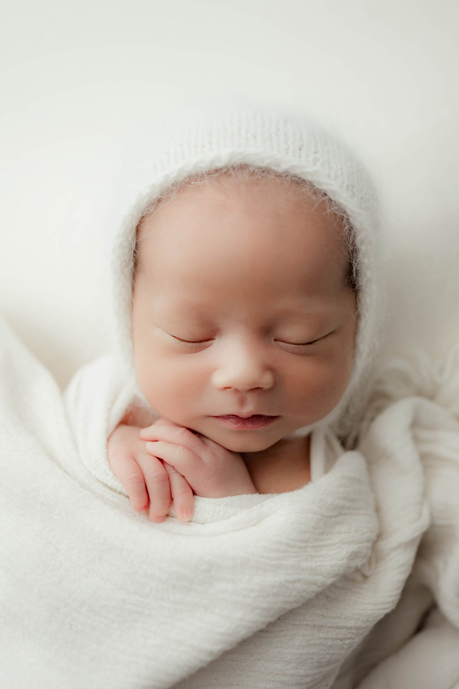 Baby boy in white hat under white blanket sleeping during newborn photo session