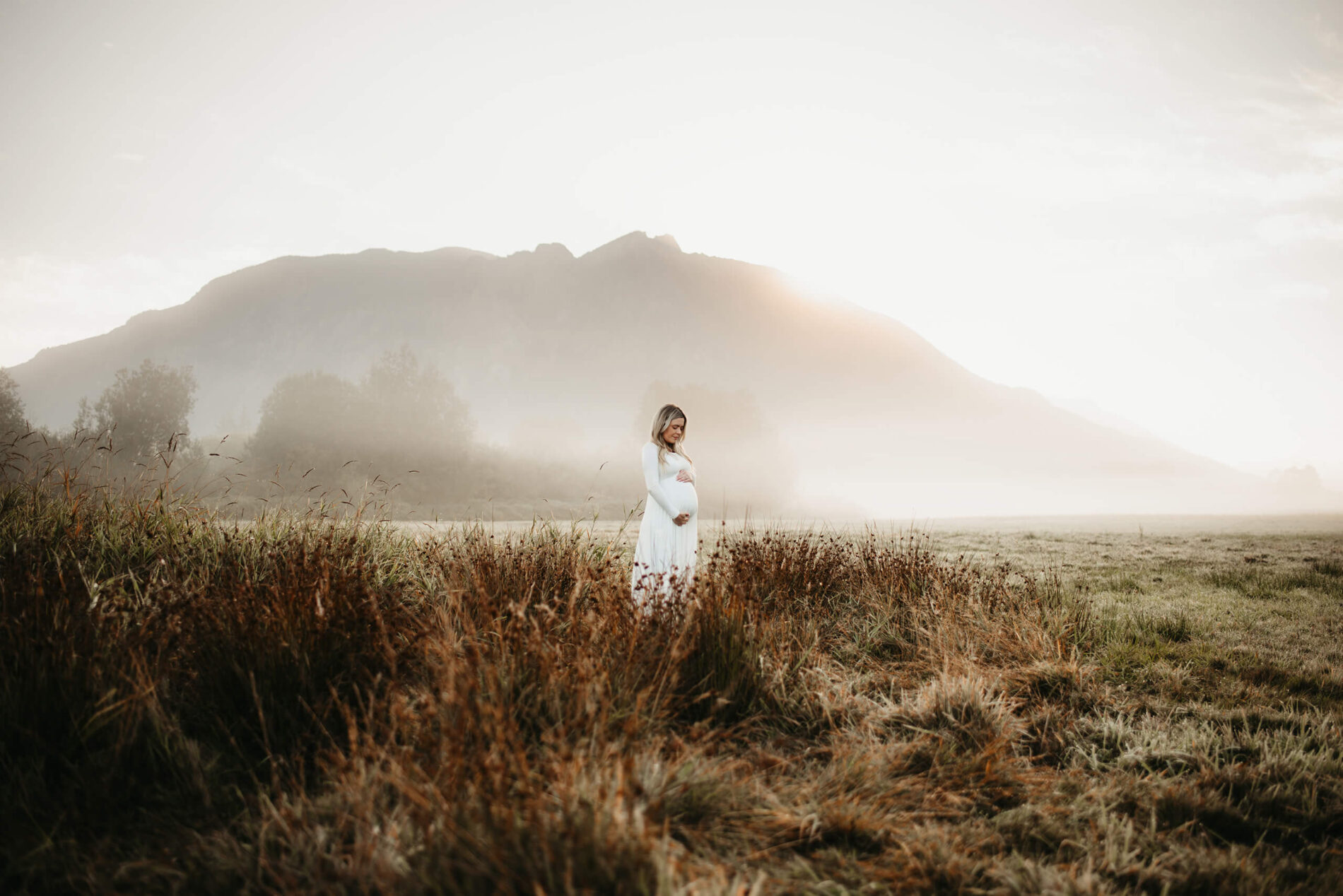 Pregnant woman during maternity photo shoot in a beautiful white dress standing a field with stunning mountain behind her during sunrise