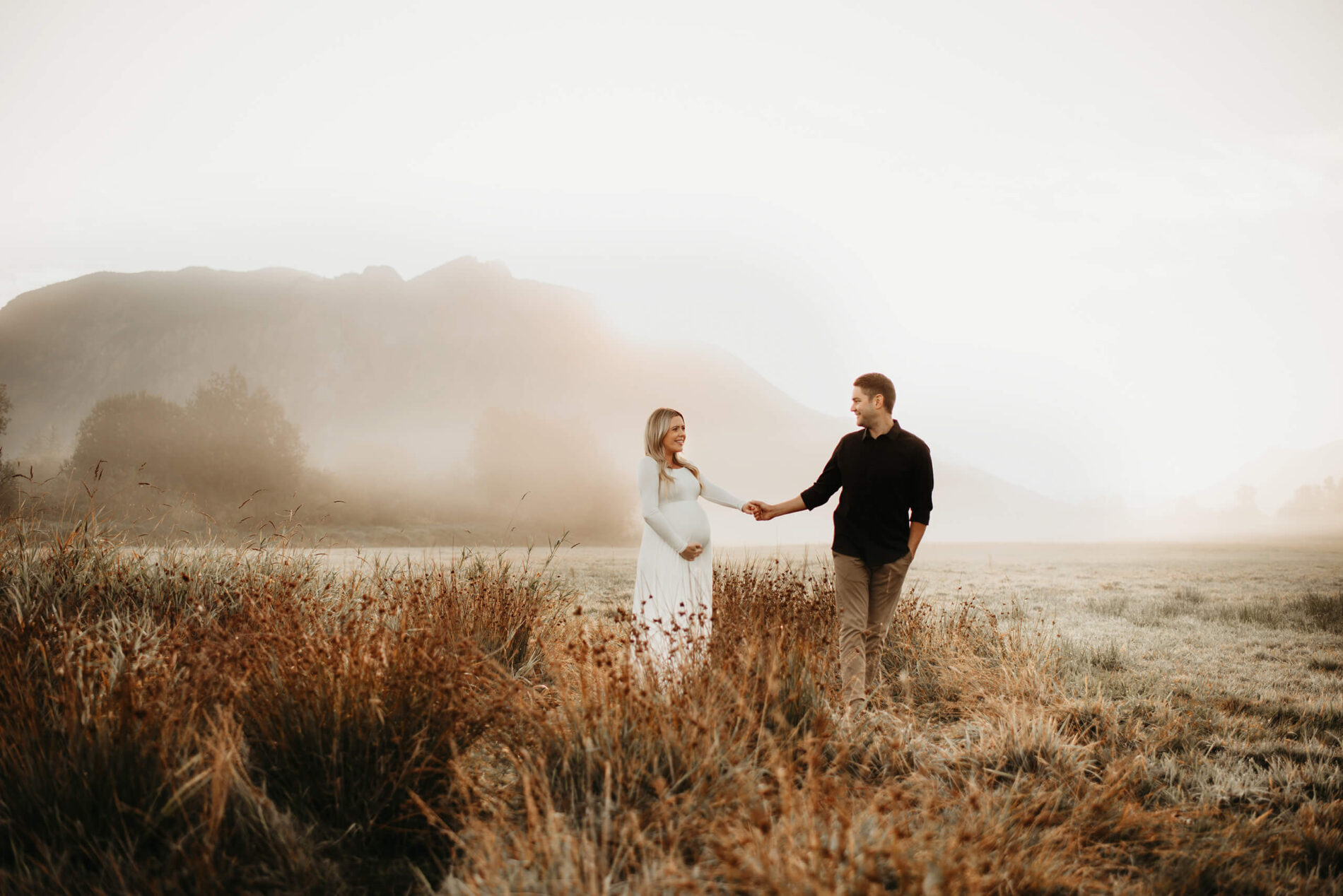 Maternity photo shoot with couple walking through a field and mountain in the background