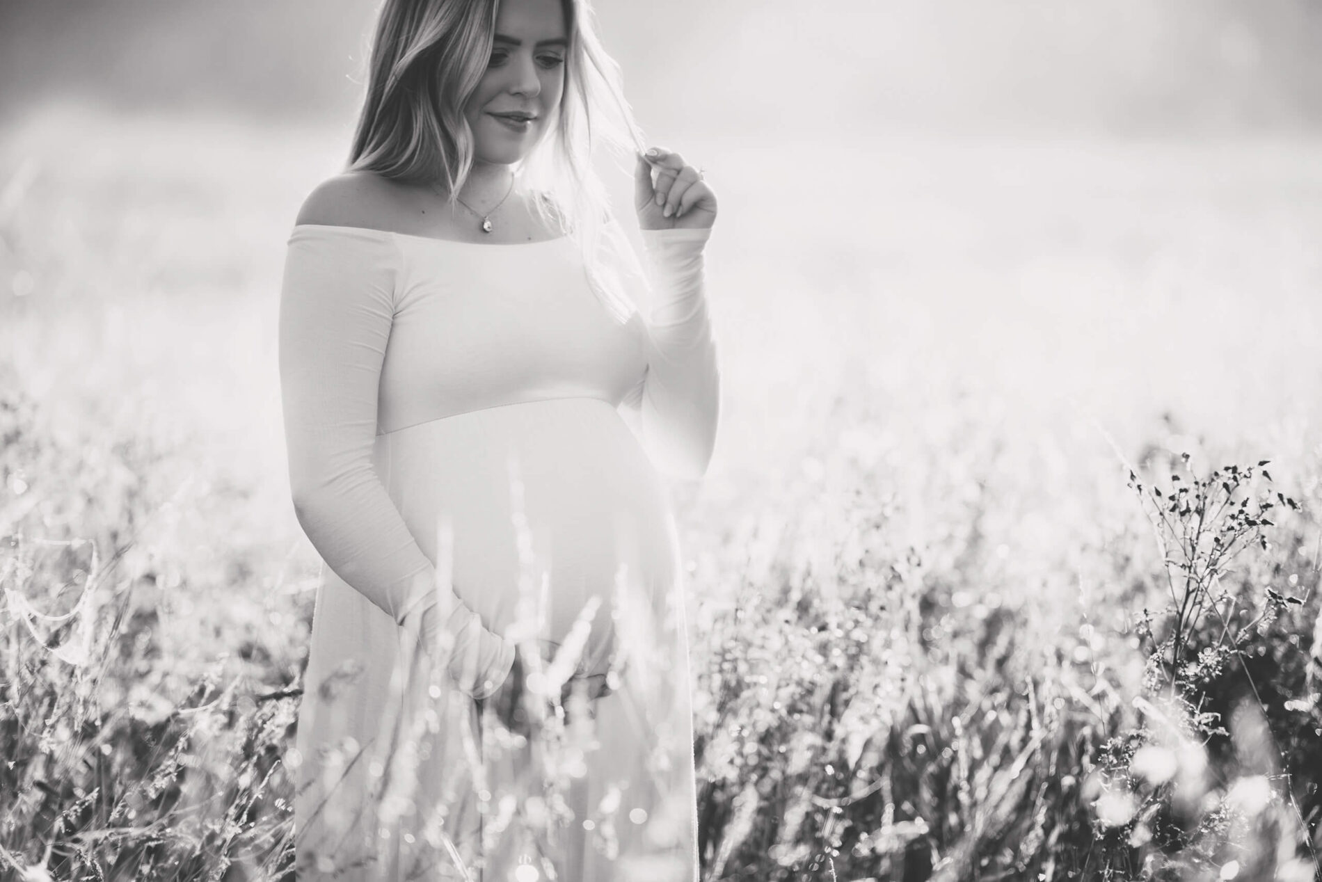 Black and white maternity photography of a happy woman in a dress standing in the middle of a field with her hand around her belly