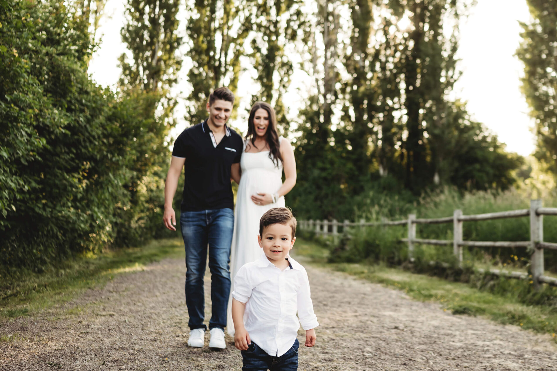 Maternity photography in the Bellevue area, smiling pregnant couple walking in a park with their toddler son in front of them and tall trees in the background