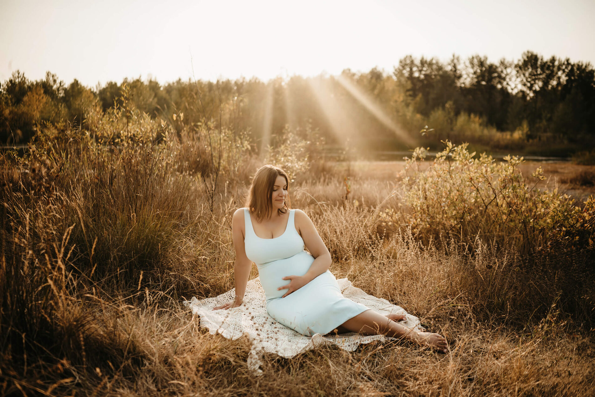 Maternity photo shoot in Bellevue area, happy woman sitting on a blanket in a field with tall dress with her hand on her belly during sunset