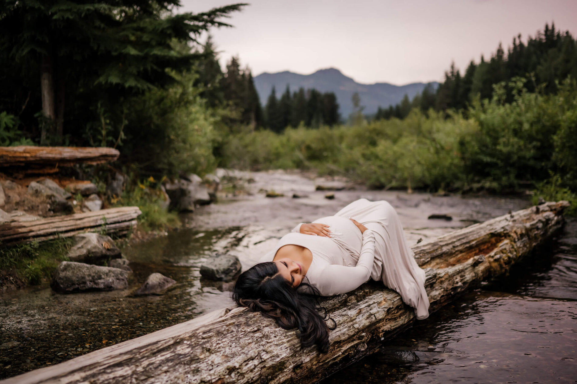 Unique maternity photography idea, pregnant woman lying on a fallen tree in a pond with mountain views