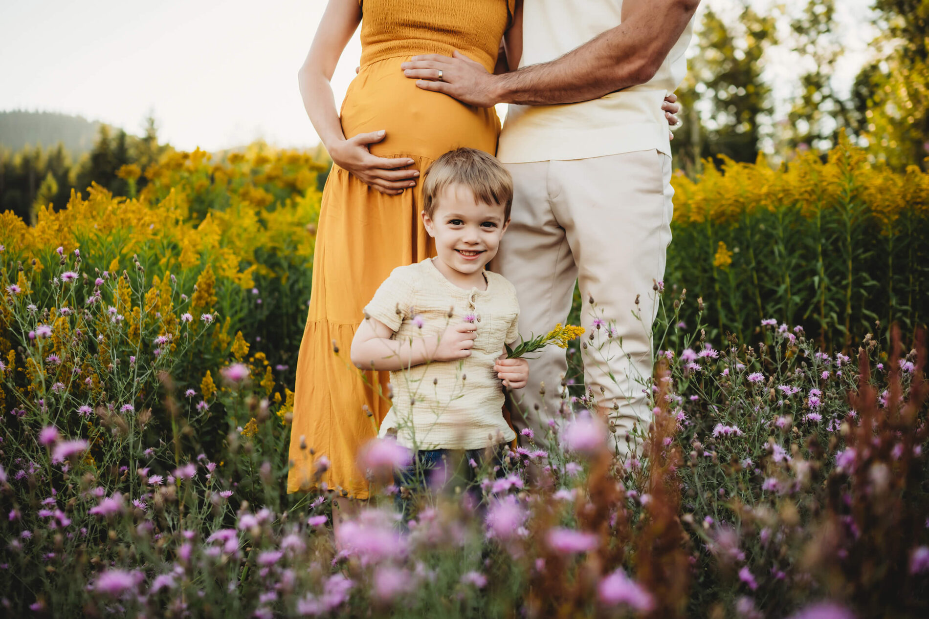 Summertime maternity photography idea with a toddler, dad's hand on mom's belly with their toddler standing in front of them