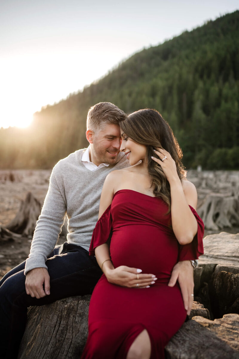 Maternity photo shoot in Seattle area, husband lovingly hugging his wife, heads lightly touching, sitting on a large tree stump with mountains in the background during sunset