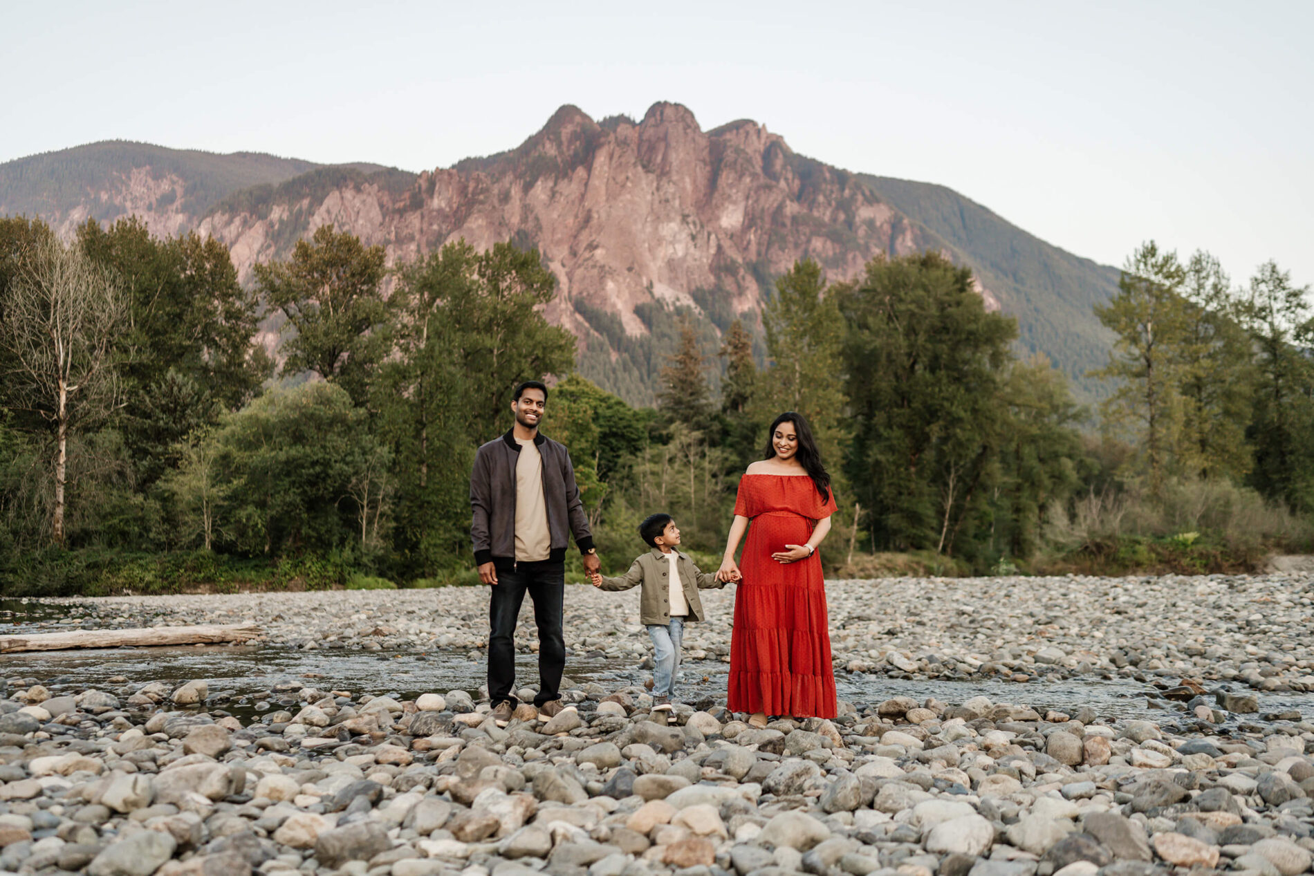 Family posing during a maternity photo shoot in Seattle on a river bank with Mount Si in the background
