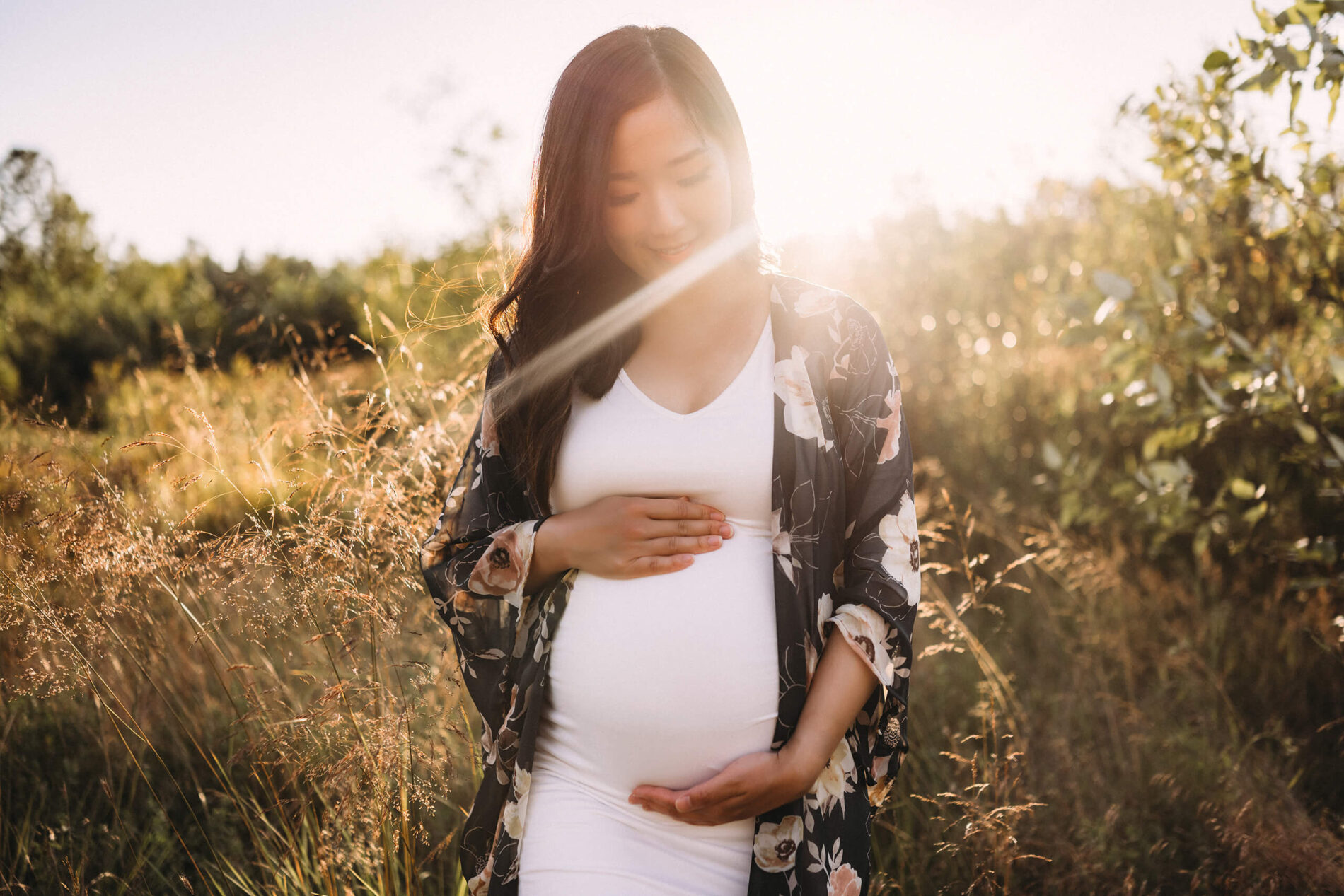 Pregnant woman posing during a maternity photo shoot, looking down at her belly, during beautiful sunset