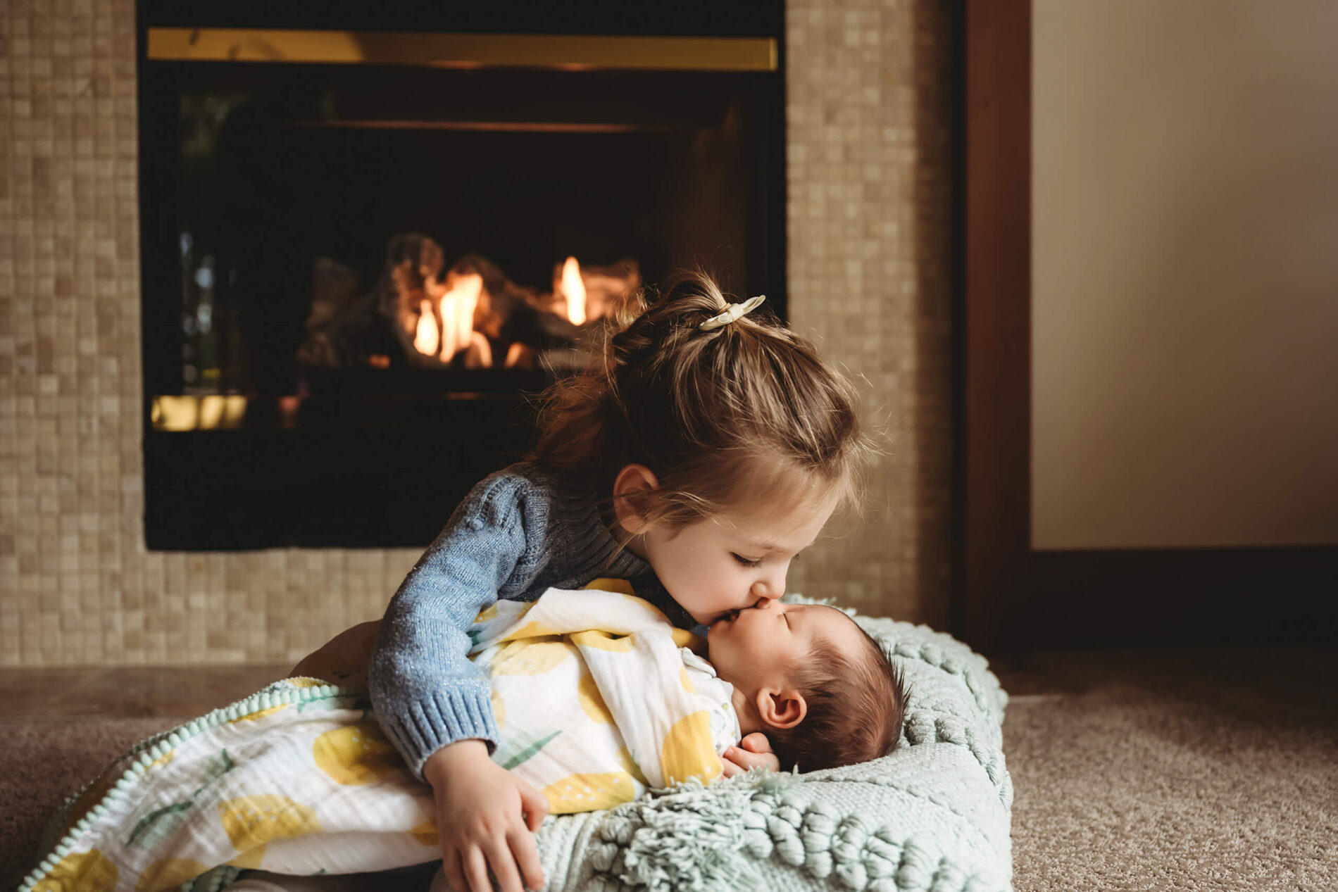 A moment during newborn photo shoot of a big sister kissing her baby sister on the floor in front of a fireplace