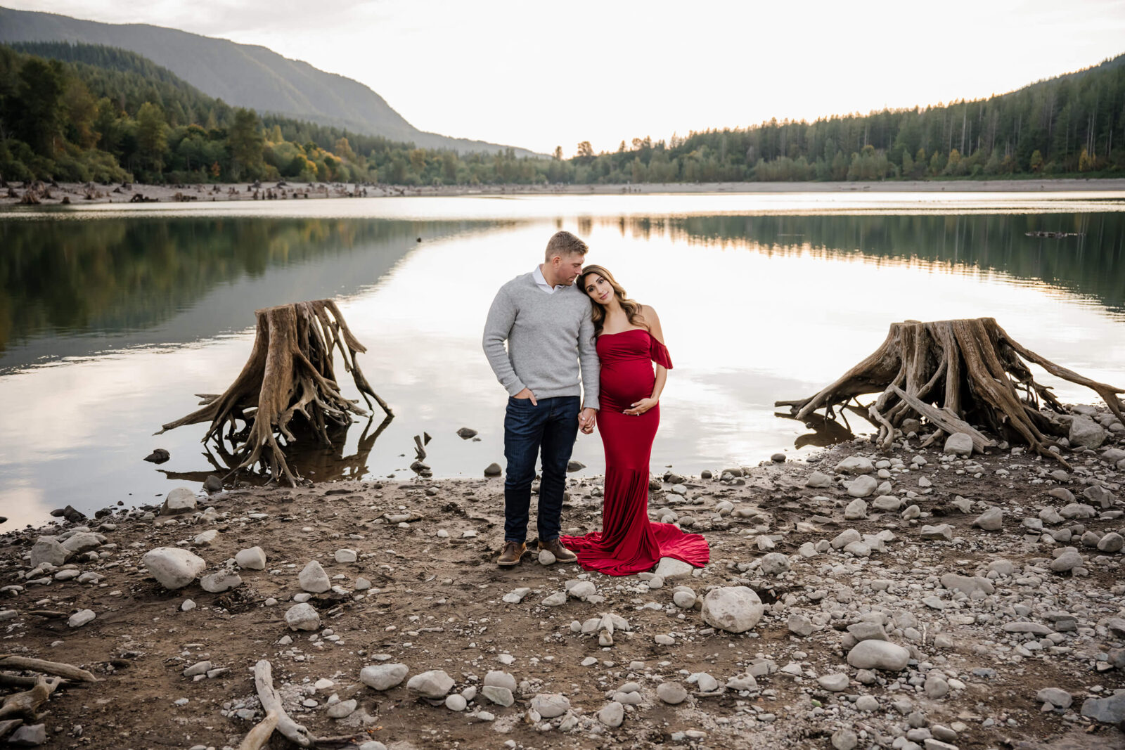 Maternity photoshoot in Seattle area at Rattlesnake Lake: expectant mom in red off-shoulder gown leans on partner as they hold hands on rocky shore; calm water mirrors Cascade evergreens at sunsethotography in Seattle, husband and wife standing on a beach of a serene lake with mountains behind them, wife leaning her head on husband's shoulder