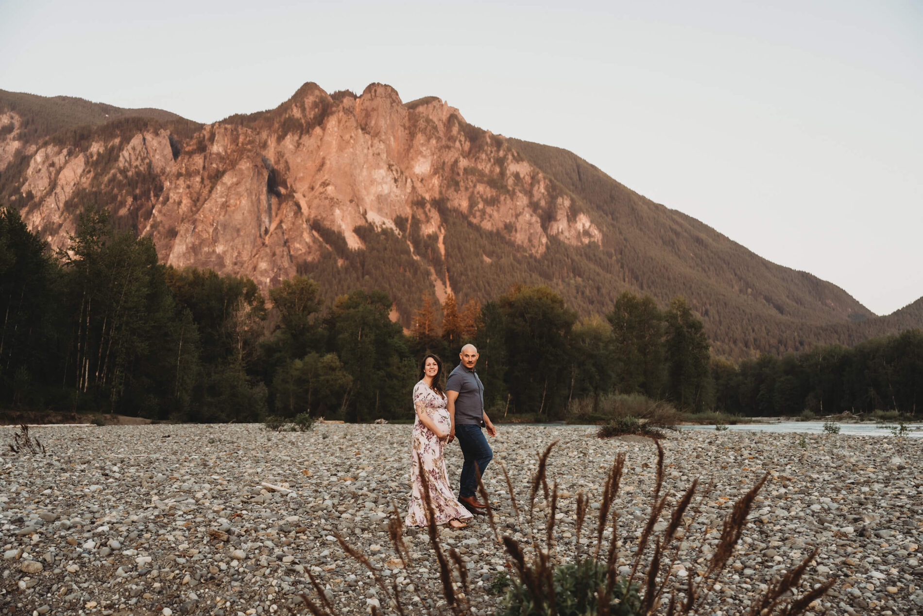 Pregnant couple walking on a riverbank during maternity photo shoot, with Cascade Mountains in the background