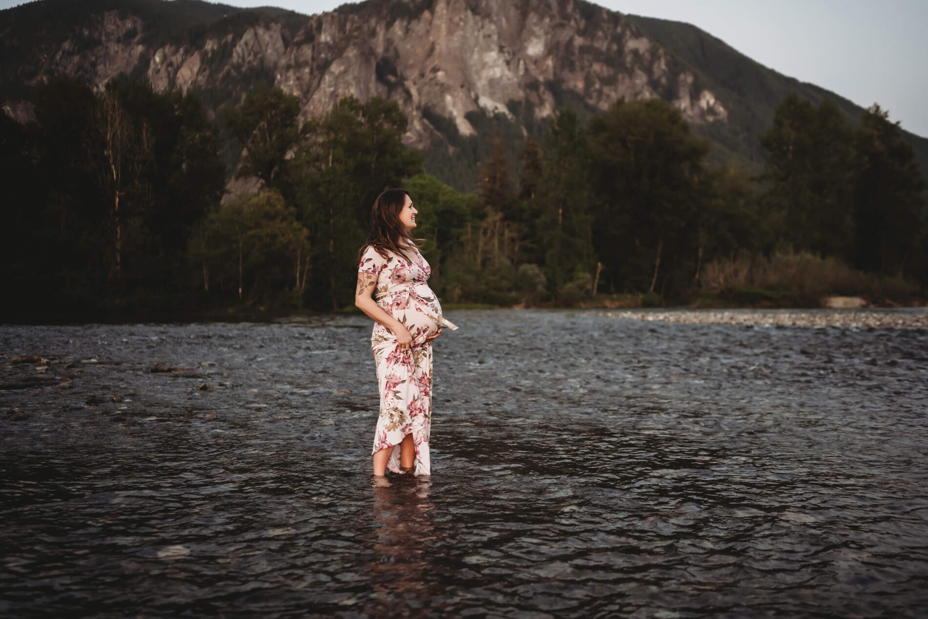 Spring pregnancy photo in Seattle sample, smiling woman in a dress standing in a river with mountains in the background