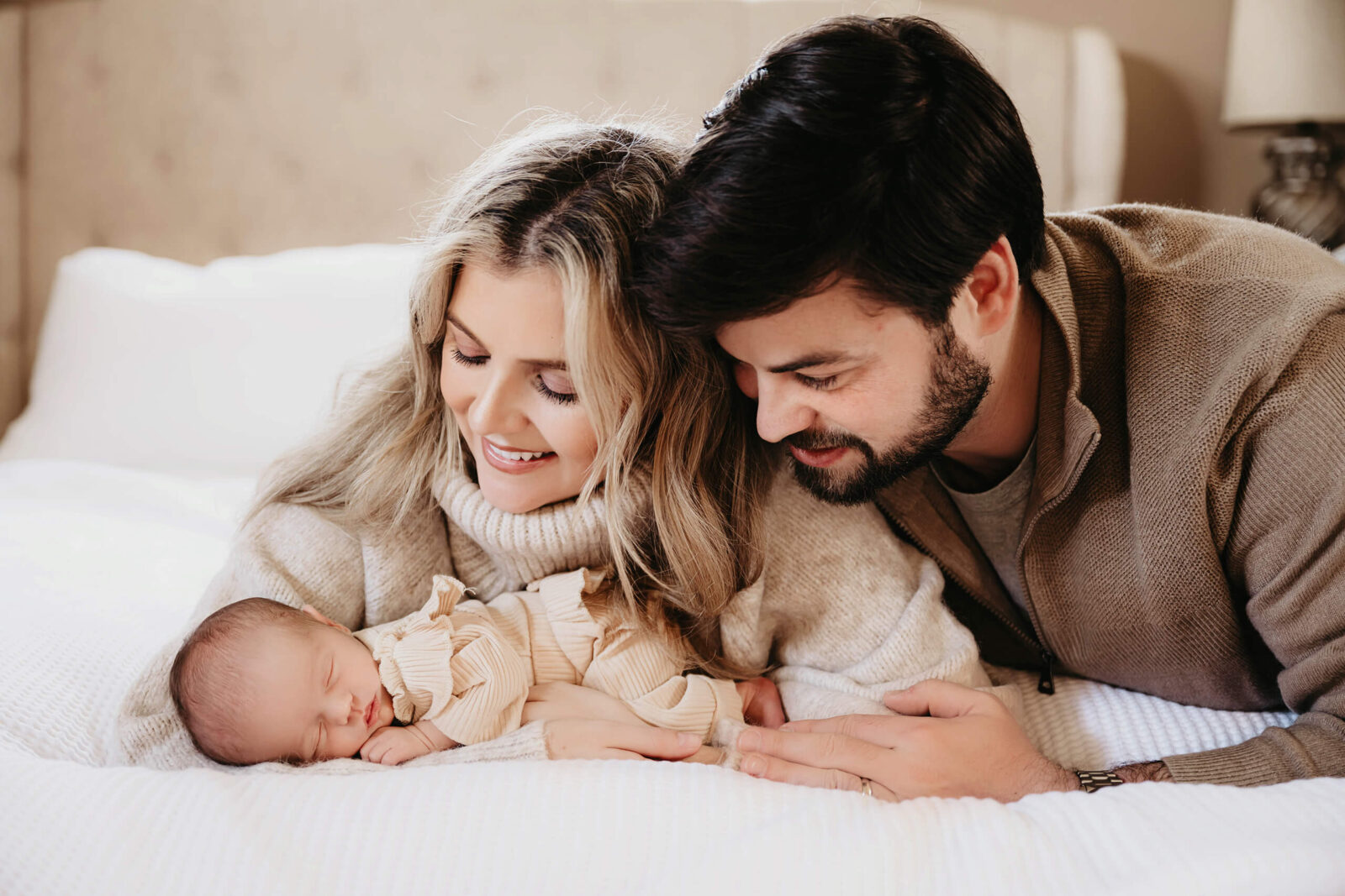 Parents cuddling close with their sleeping newborn on a bed during an in-home newborn photo session
