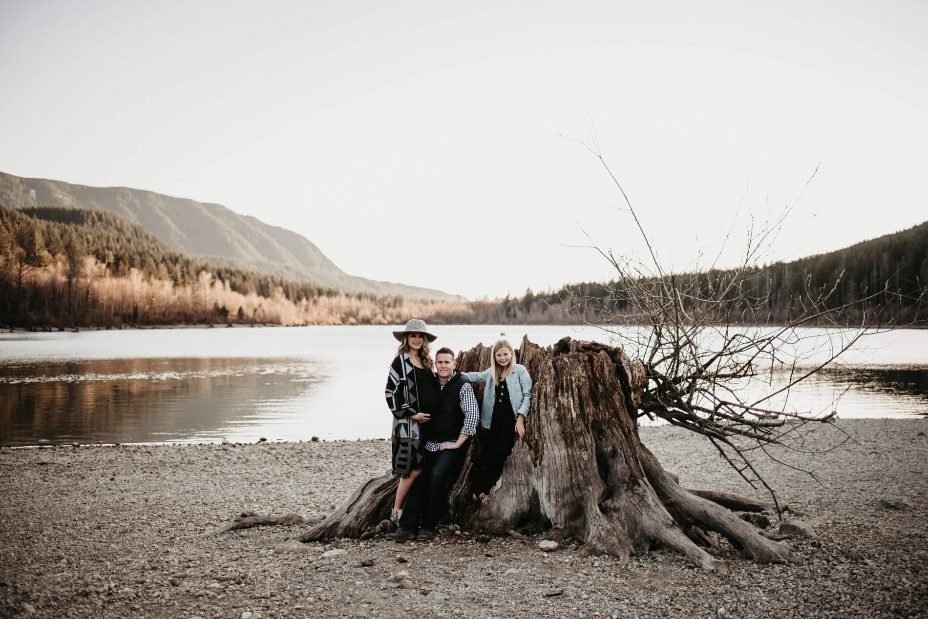 Pregnancy photo shoot, mom, dad and young girl posing next to a big tree trunk with lake and mountain views