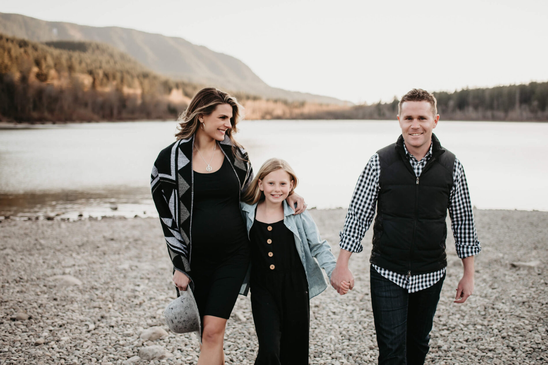 Family shot during a maternity photo session, dad, mom, and young girl holding hands on a beach of a lake