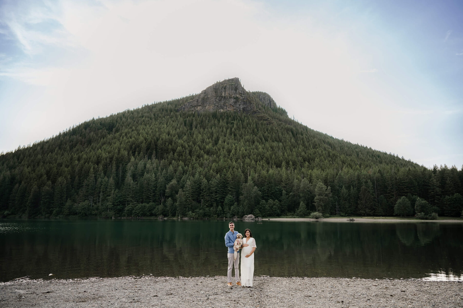 A couple standing on a beach of a lake during maternity photo shoot in Seattle with mountains in the background