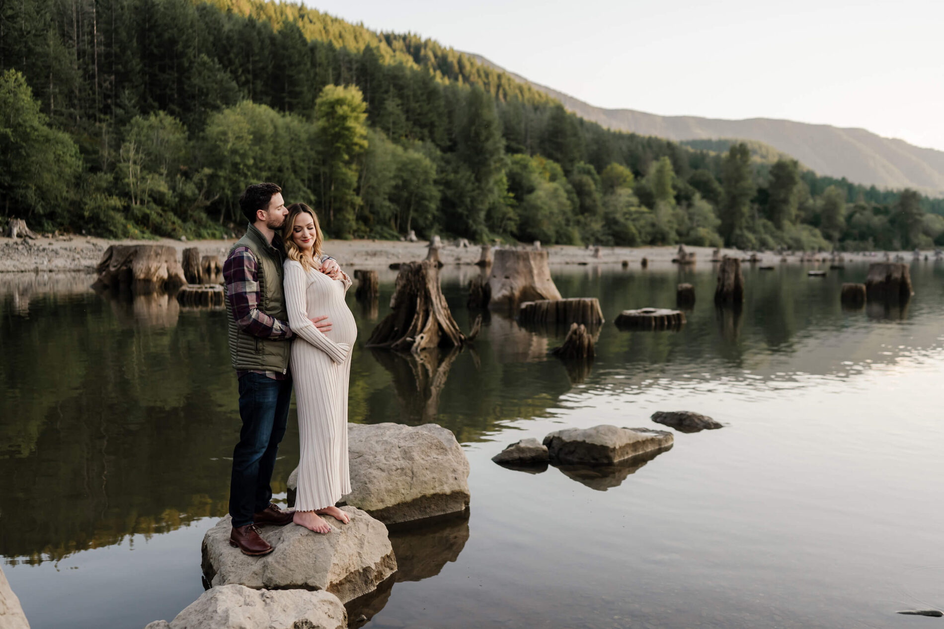 A couple posed on a rock in a lake during a maternity photo shoot in Seattle area, Cascade mountains in the background
