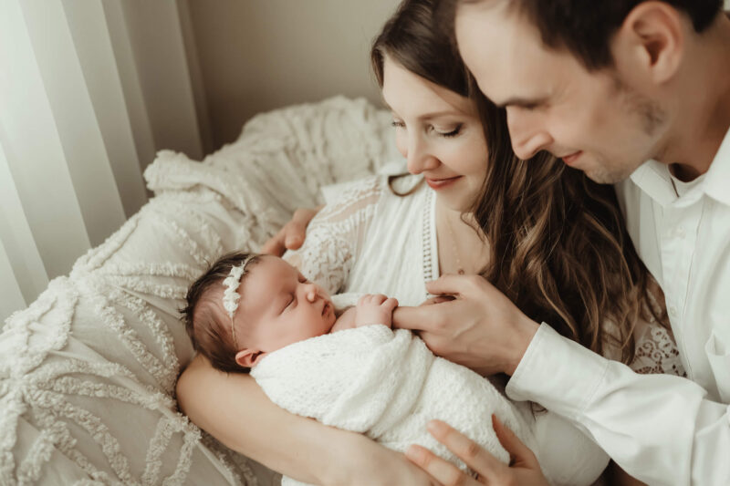 Mom cradles a sleeping newborn wrapped in white while dad leans close and touches the baby’s hand.