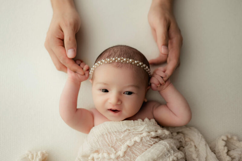Dad’s hands gently hold both hands of an awake newborn wearing a pearl headband.