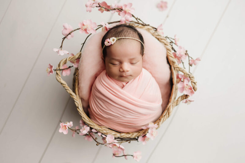 Sleeping newborn wrapped in pink in a woven basket surrounded by pink blossoms.