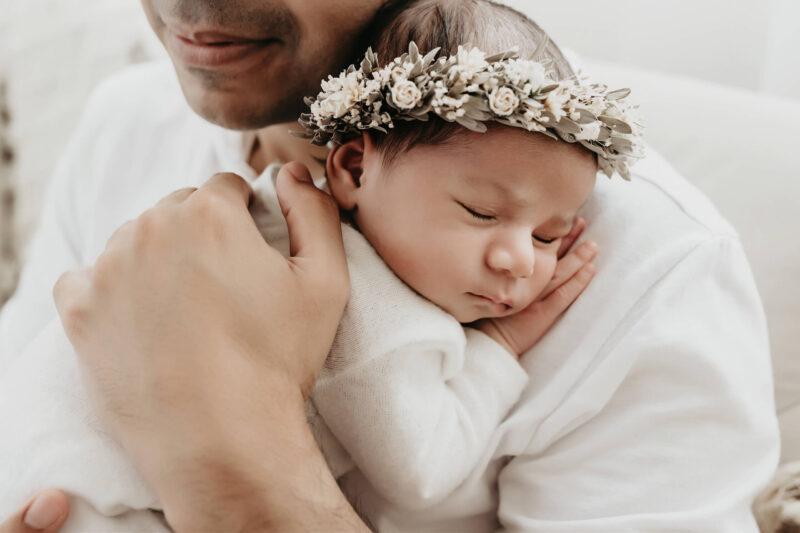 Dad holds a sleeping newborn wearing a floral crown against his chest.