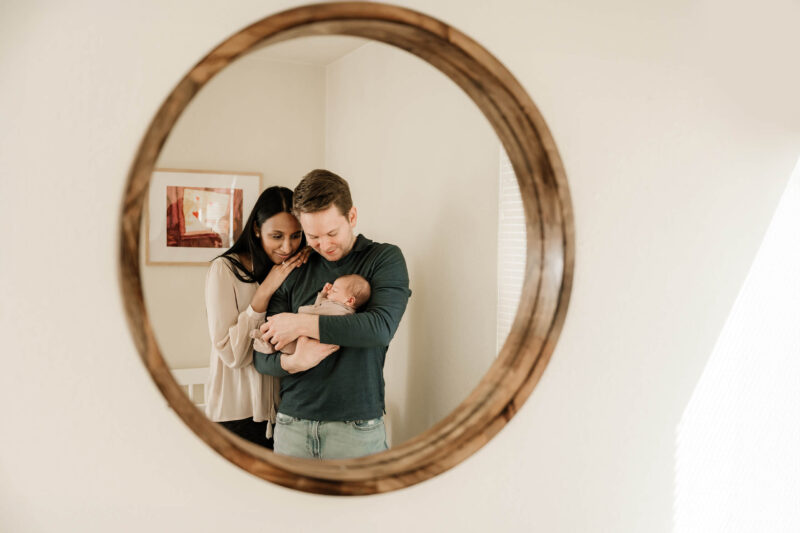Mom leans close as dad cradles their newborn, reflected in a round wall mirror.