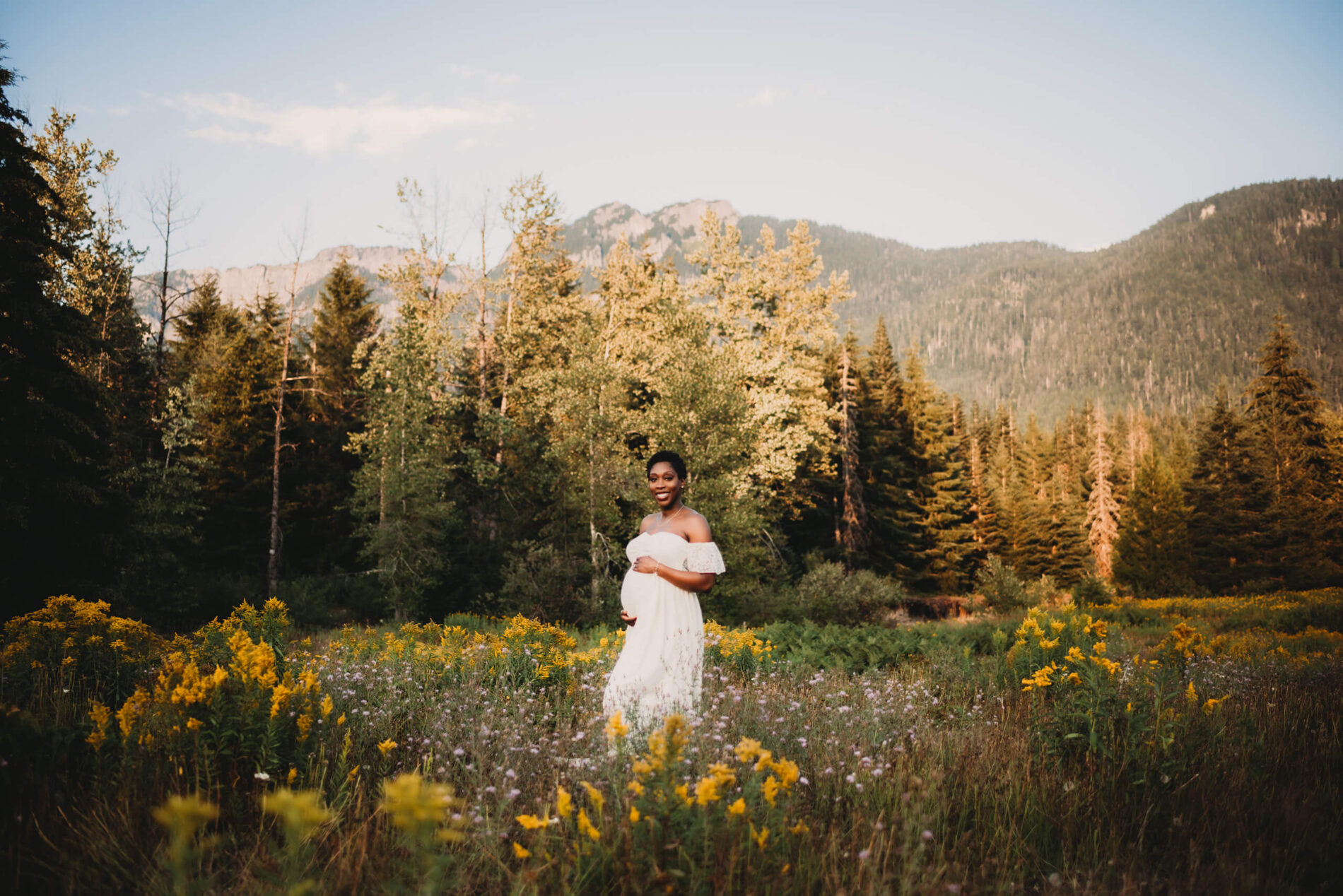 A smiling pregnant woman in a white dress in a field of wildflowers during a Seattle maternity photoshoot