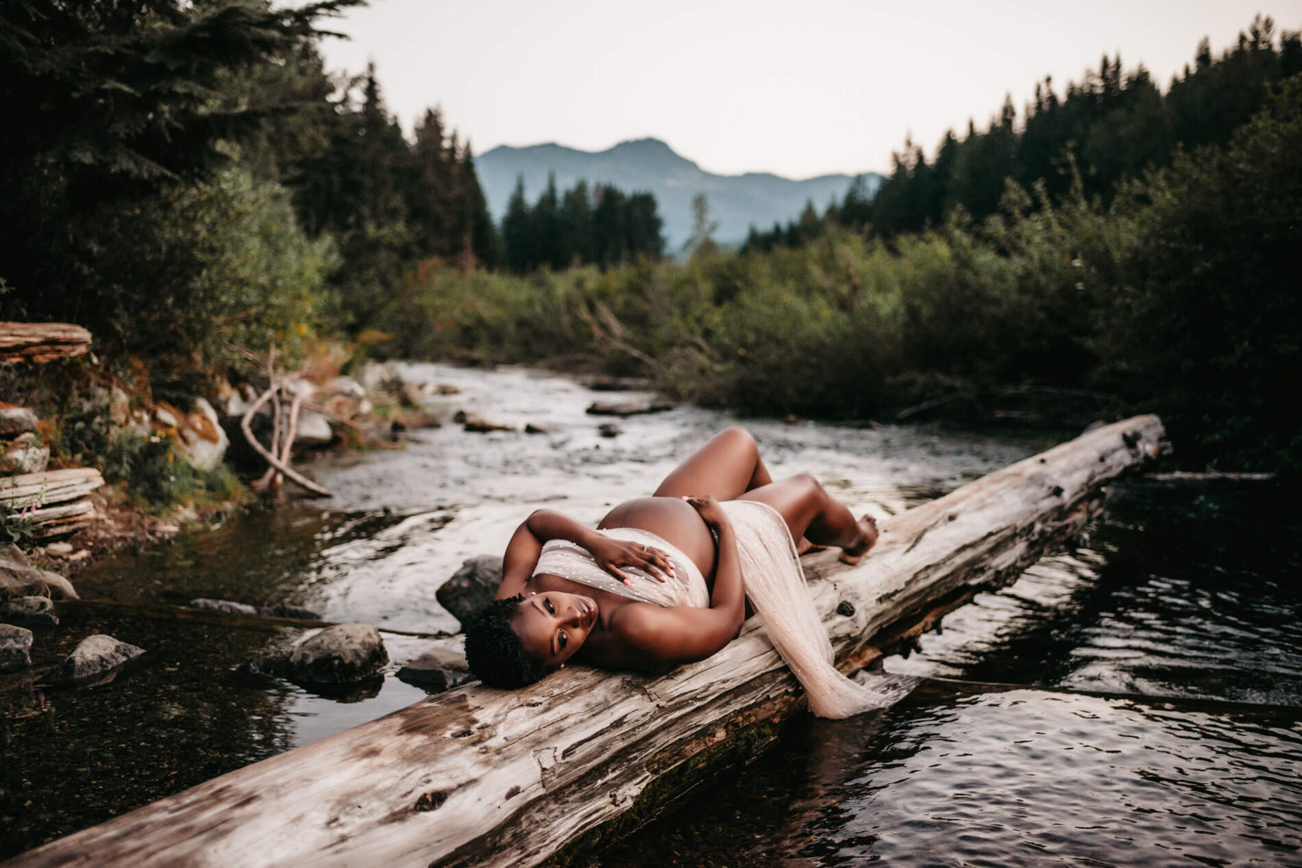 Seattle maternity photography with mountain backdrop, young woman posed on a log over running water