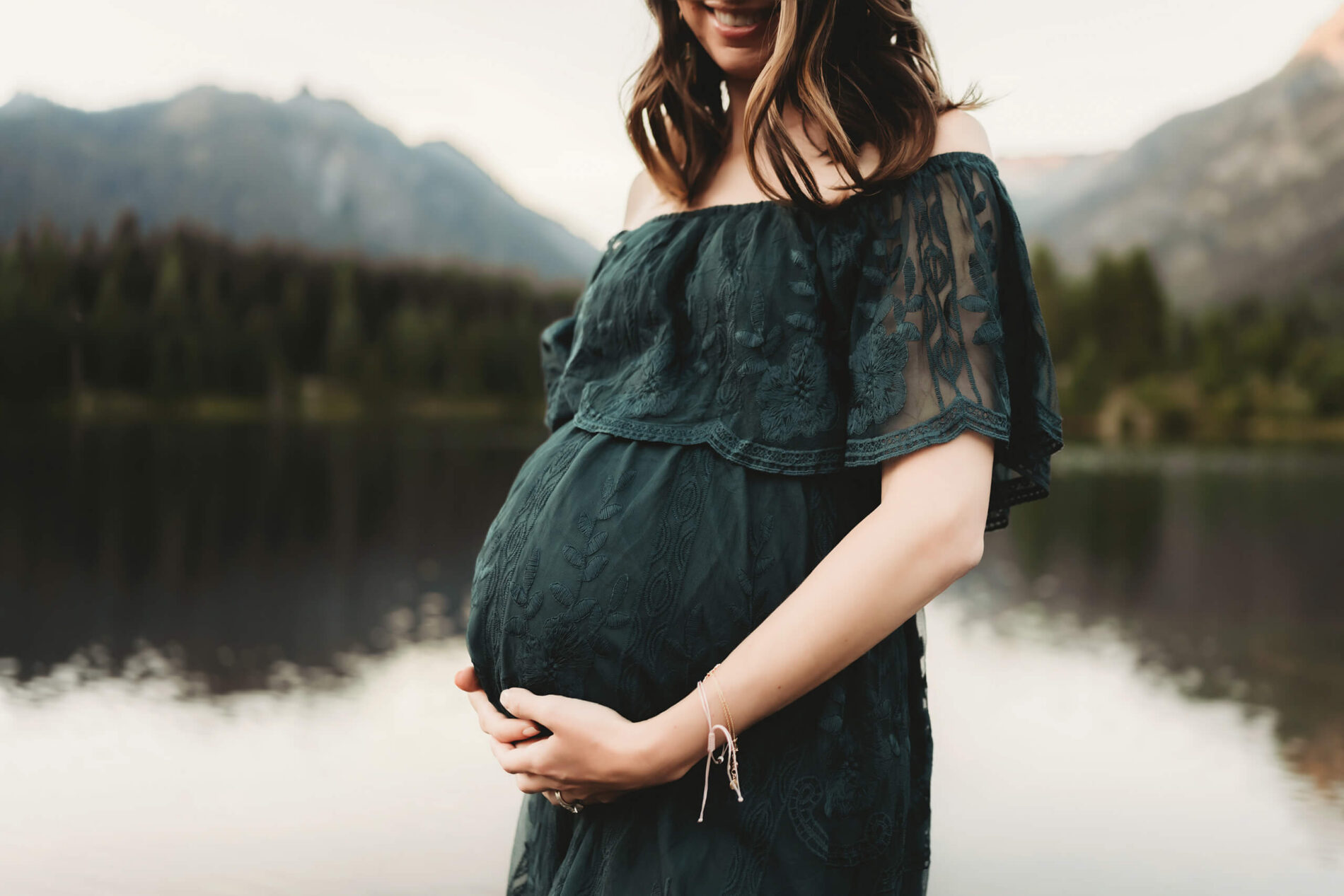 Posing idea for pregnancy photos in Seattle, mom in green dress holding her belly, pond and mountains in the background